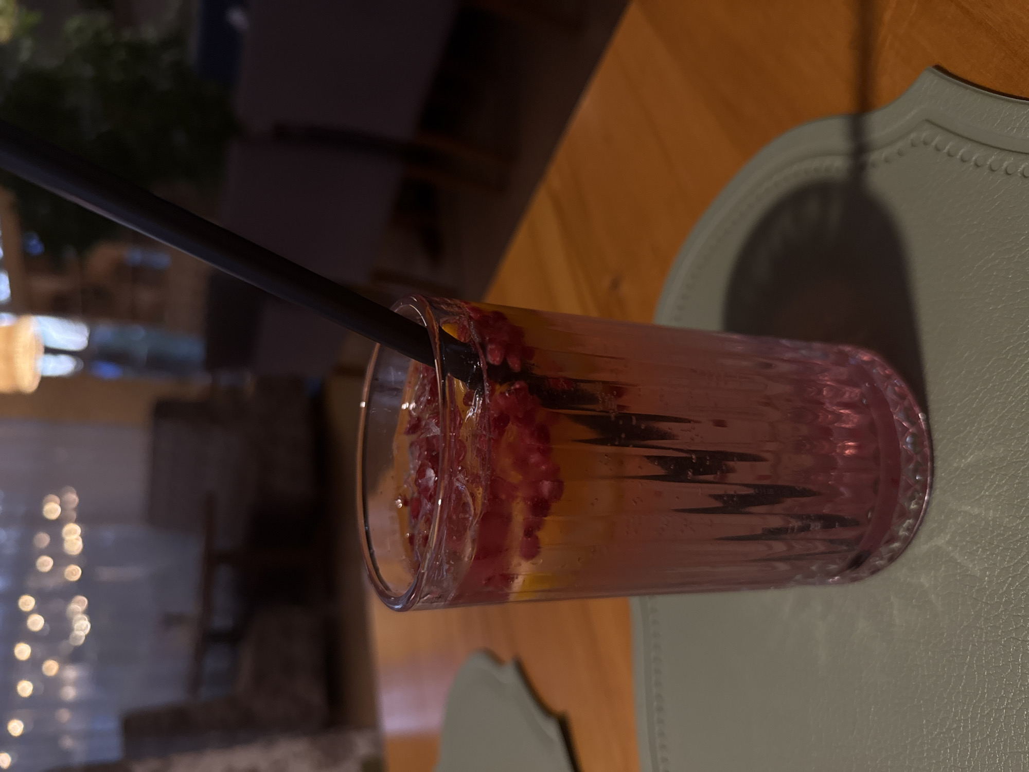 A close-up, eye-level, indoor shot captures a tall, fluted glass filled with a pink beverage containing ice, citrus slices, and red berries. A black straw extends from the top of the drink. The glass rests on a textured, light green placemat, which is on a wooden table. In the background, out of focus, is a dimly lit restaurant interior with upholstered chairs and tables, and string lights hanging from the ceiling. The setting is likely a cafe or restaurant in Abinsk, Russia, given the provided context.