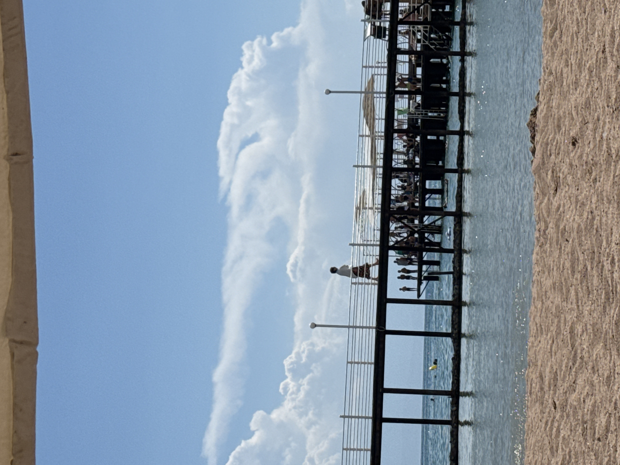 The image captures a bright, sunny day at a beach resort. In the foreground, a sandy beach transitions to clear, shallow water. Beyond the water, a wooden pier stretches out over the sea. The pier is crowded with people, some are walking, while others are sitting or standing, enjoying the view. The pier has a metal railing and several straw umbrellas providing shade. A single person is walking on the pier, silhouetted against the bright sky. The sky is a vibrant blue with fluffy white clouds. The location is identified as Sary-Oy, Kyrgyzstan, a resort town on the northern shore of Issyk-Kul Lake. The overall scene depicts a lively and recreational atmosphere at a lakeside resort.