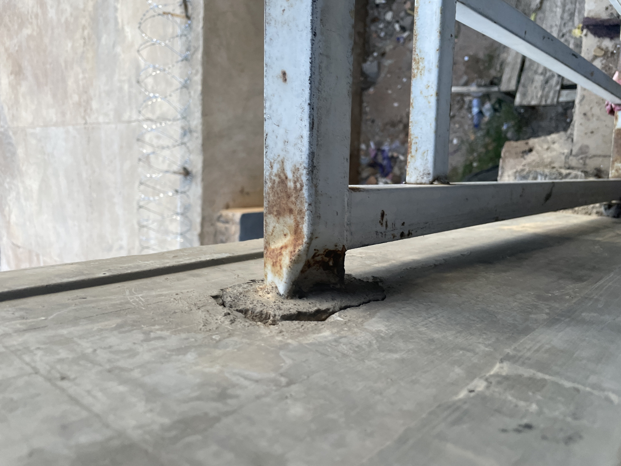 The image shows a close-up view of a rusted metal railing attached to a concrete wall. The railing is light blue with significant areas of rust, particularly at the attachment point to the wall. The concrete wall appears weathered and has a chipped section around the railing's anchor. In the background, out of focus, is a glimpse of barbed wire strung along what appears to be a perimeter. Below the railing, a messy, somewhat debris-strewn area is visible, suggesting an outdoor, possibly neglected urban environment. The scene is likely located in the city of Jalingo, Nigeria, based on the contextual information provided.