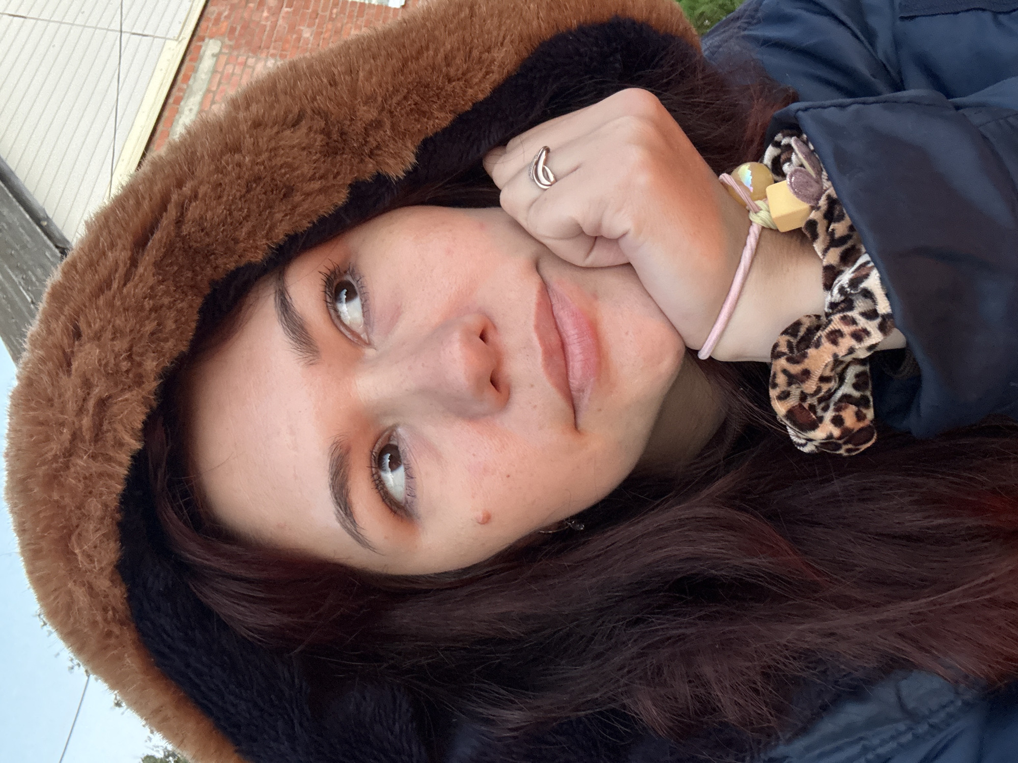 A close-up, eye-level, outdoor medium shot features a young adult with long, dark auburn hair wearing a dark blue hooded jacket with a brown faux fur lining. The subject's right hand is resting on their chin, and they are wearing a ring on their right index finger. Two scrunchies are visible on the subject's left wrist. The background includes a brick building and a light-colored structure, likely part of a house, with a clear sky above. The lighting suggests it is daytime. The image does not depict Imelevo, Russia.