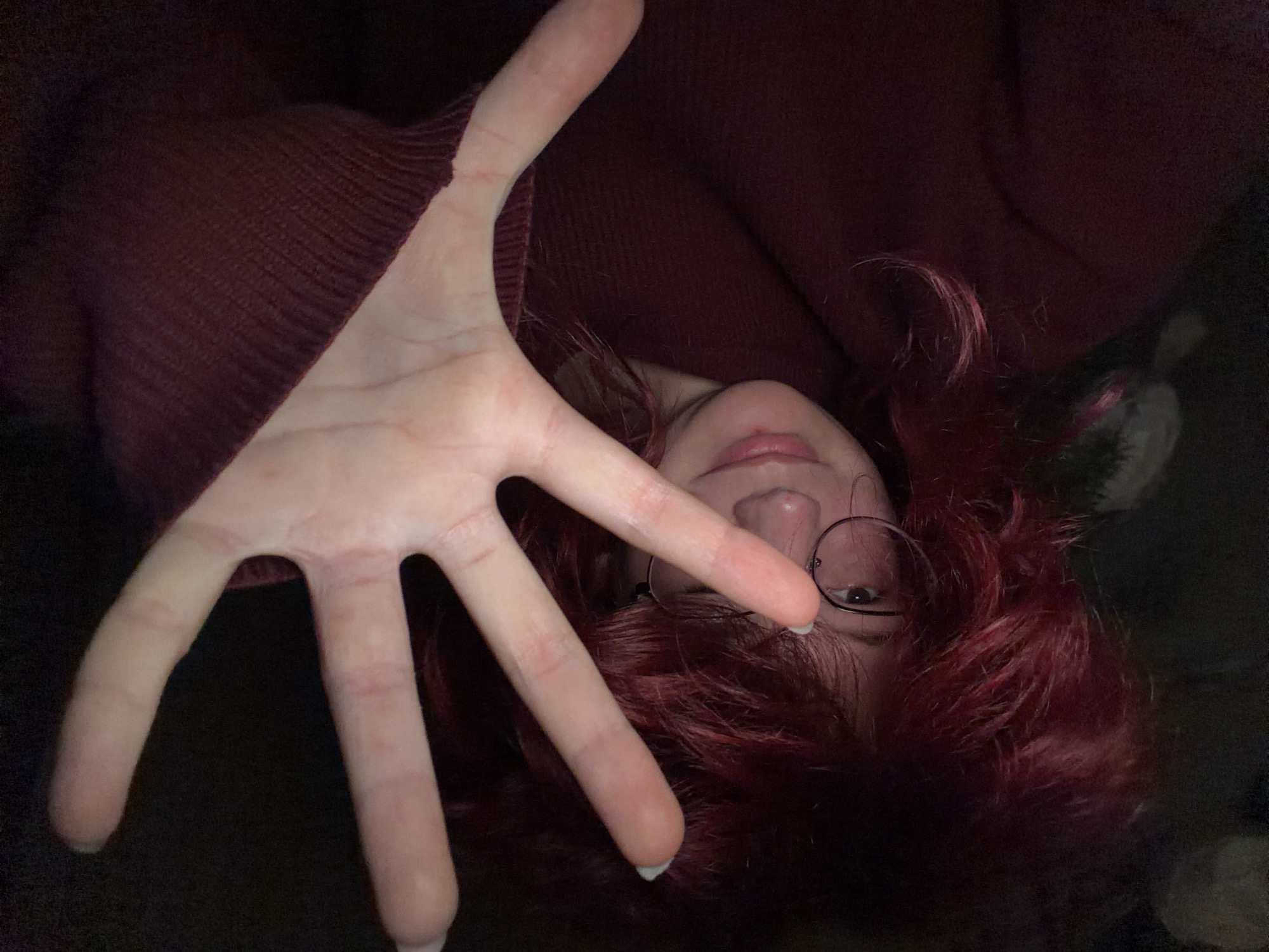 The image is a selfie taken indoors in low light. A person with vibrant red, voluminous hair and glasses is partially visible on the left side of the frame. Their right hand is held up towards the camera, obscuring a portion of their face. The person is wearing a dark red, textured sweater. The background is dark and indistinct. There is no information in the image to suggest a specific location within Chelyabinsk, Russia.