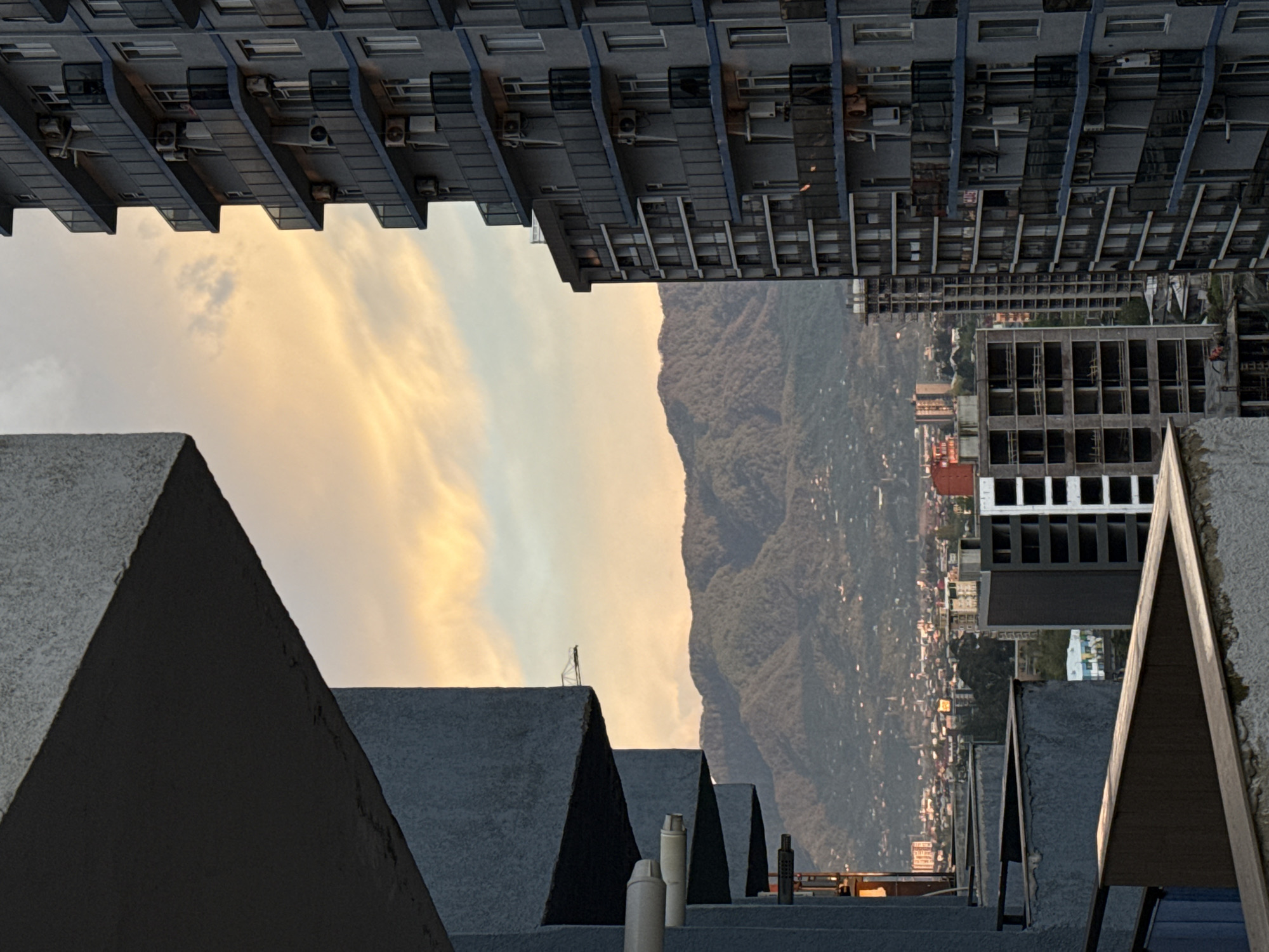 The image shows an upside-down view of a landscape in Batumi, Georgia. Above, there are multiple rows of high-rise buildings with a lot of air conditioning units outside of the windows. In the center of the image, there is a mountain range and the outskirts of the city, a lower built cityscape. In the foreground, there are a series of angled rooftop structures, possibly the roofs of several buildings, with some piping extending from the roofs. The sky is partially visible, with light-yellow, cloud filled sky filling the gaps between the buildings and mountain in the background.