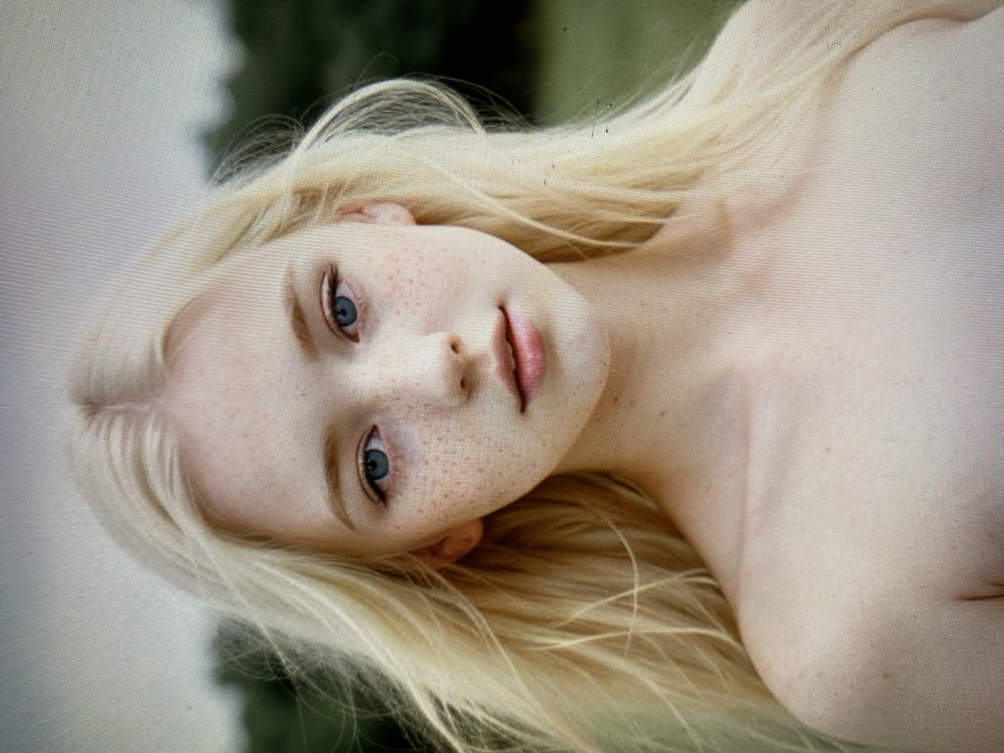 A close-up, eye-level, medium shot of a young Caucasian female with fair skin and a smattering of freckles across her nose and cheeks. Her long, blonde hair is windswept and frames her face. She has bright blue eyes and her lips are slightly parted. Her shoulders and upper chest are bare and visible. The background is blurred, with dark green foliage suggesting an outdoor setting, possibly a park or a wooded area. The lighting is soft and natural. The scene is static, with no apparent action taking place. The image appears to be a portrait.