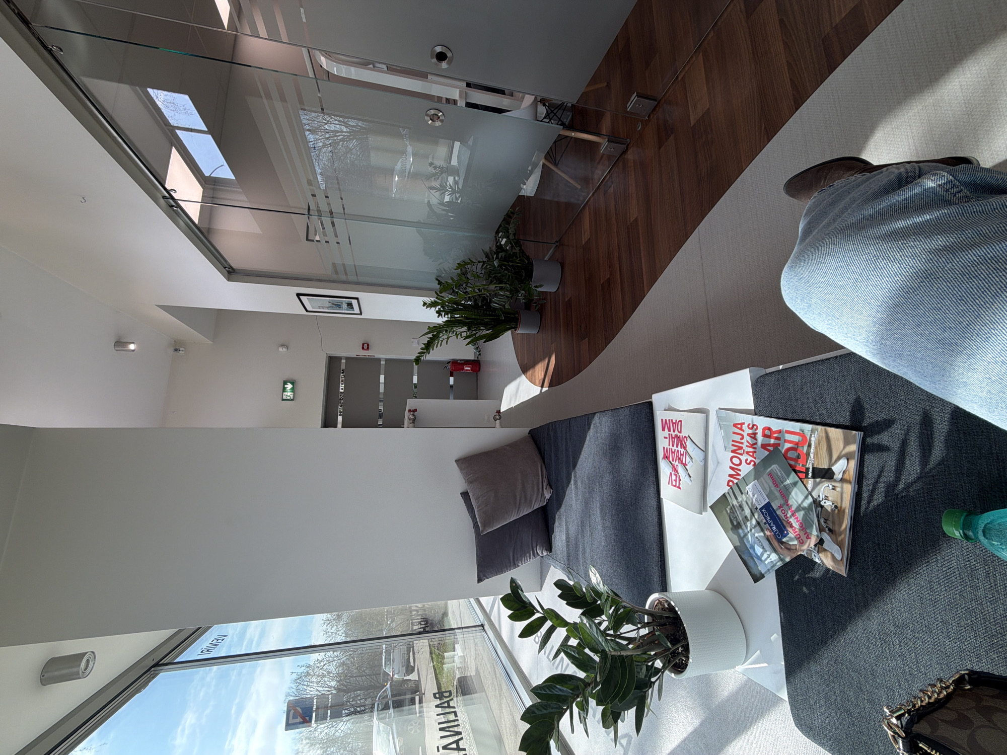 This image depicts a bright and modern waiting area, likely in a dental or medical clinic, filled with natural daylight. A person is seen from the waist down, wearing blue jeans and dark shoes, seated on a dark grey upholstered sofa on the right side of the frame. Adjacent to them, on a low white table, sits a potted ZZ plant, several magazines, a green-capped bottle, and a dark handbag with a gold chain strap. Notable text on the magazines includes "CURAPROX ALIGNER foam 40ml" and "MONIJA SĀKAS AR," pointing towards a dental or orthodontic context.
To the left, a long, dark grey upholstered bench with two grey cushions runs along a white wall. Large windows on this wall look out onto a street, revealing cars and bare trees under a clear sky, indicating a sunny day during a cooler season. The word "BALTIJAS" is faintly visible in reflection on the window. Further back, glass partitions with frosted stripes lead to an interior corridor. Within this corridor, a red fire extinguisher, an emergency exit sign, and a sign above a door reading "INFEKCIOZIE ATKRITUMI" (Infectious Waste) and "ORTODONTIJA" (Orthodontics) are discernible, reinforcing the clinical setting. The flooring transitions from a light, textured material to a warm, wood-patterned surface. The overall environment is clean, professional, and well-lit, consistent with a facility in Riga, Latvia.