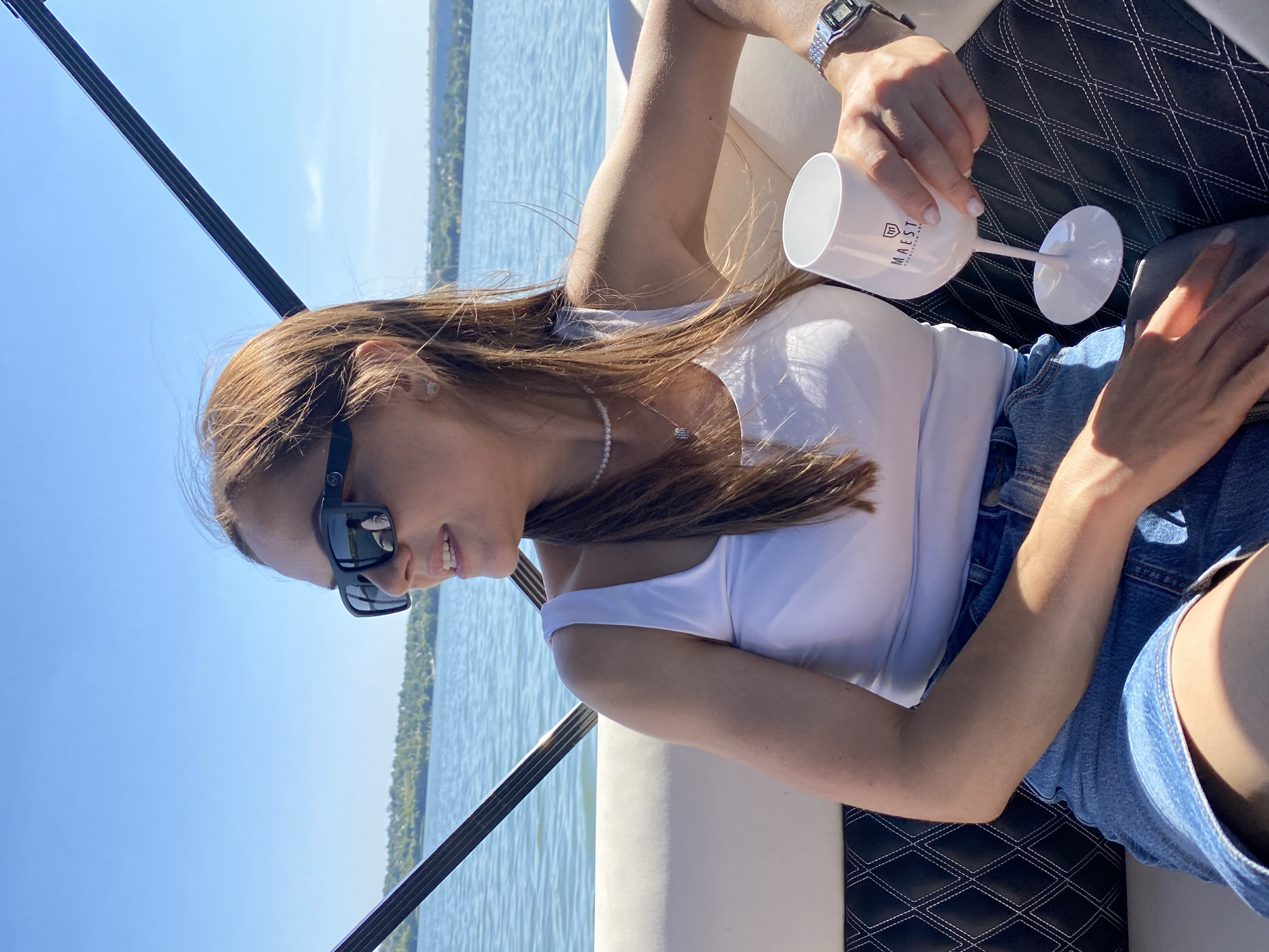 A medium shot captures a woman sitting on a boat on a sunny day. She is wearing a white tank top, denim shorts, sunglasses, a pearl necklace, and a watch. She is holding a white wine glass with the "MAESTRO ENOLOGIA ART" logo on it. The boat is on a body of water, with a tree-lined shore in the background. The sky is clear and blue. The location is the city of Molovata, Moldova.