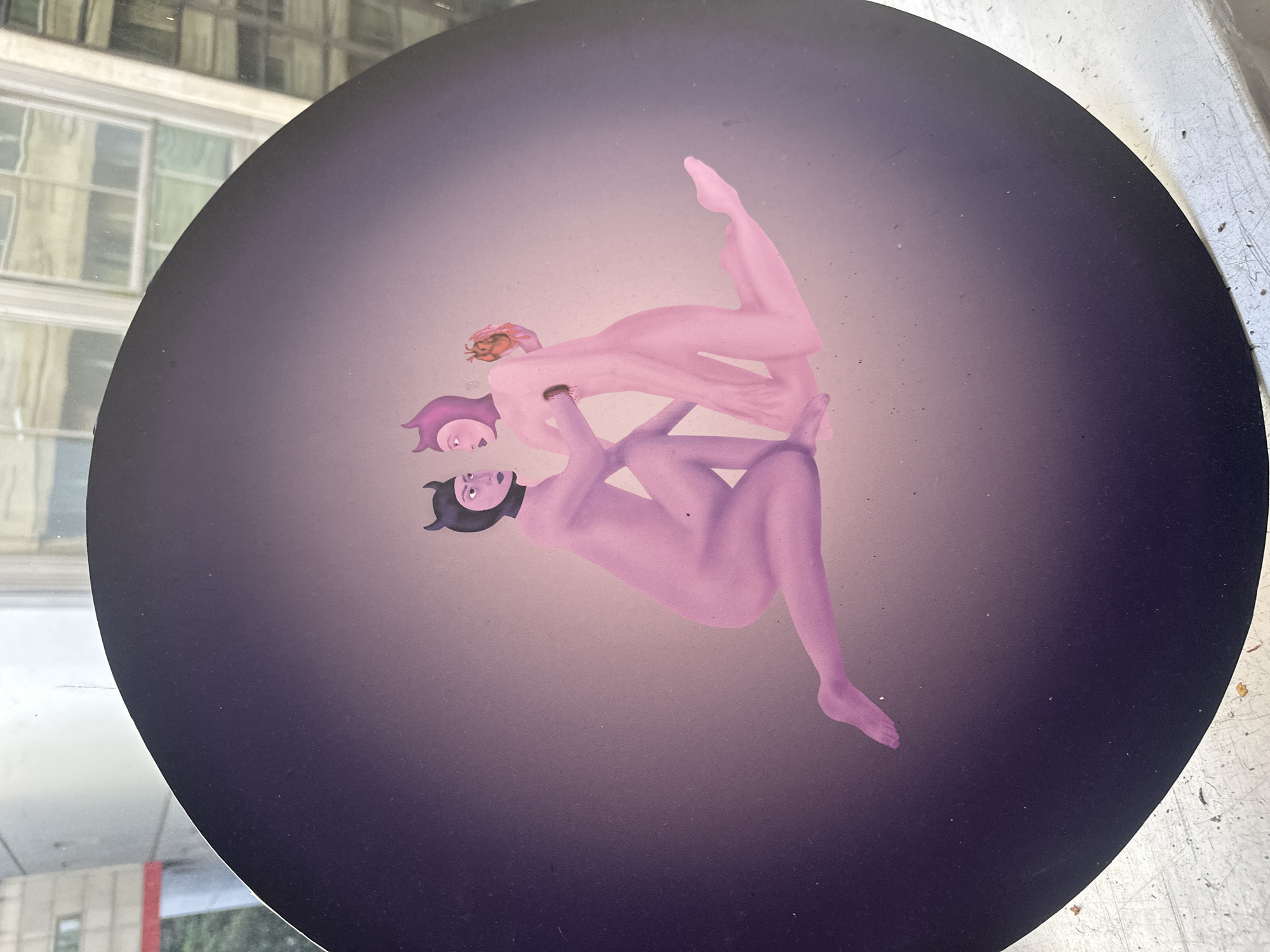 The image shows a circular artwork featuring three stylized, nude figures in shades of pink and purple. The figures are arranged in a stacked, almost acrobatic pose. One figure at the bottom appears to wear a black horned helmet or head covering. The figure in the middle wears a purple cap. The top figure has reddish-brown hair. The background is a gradient of light to dark purple. The artwork is located in the city of Buenos Aires, Argentina. The artwork seems to be located on or in front of a window, reflecting the exterior buildings of the city. The surface it is sitting on appears to be worn and light-colored, possibly metal.