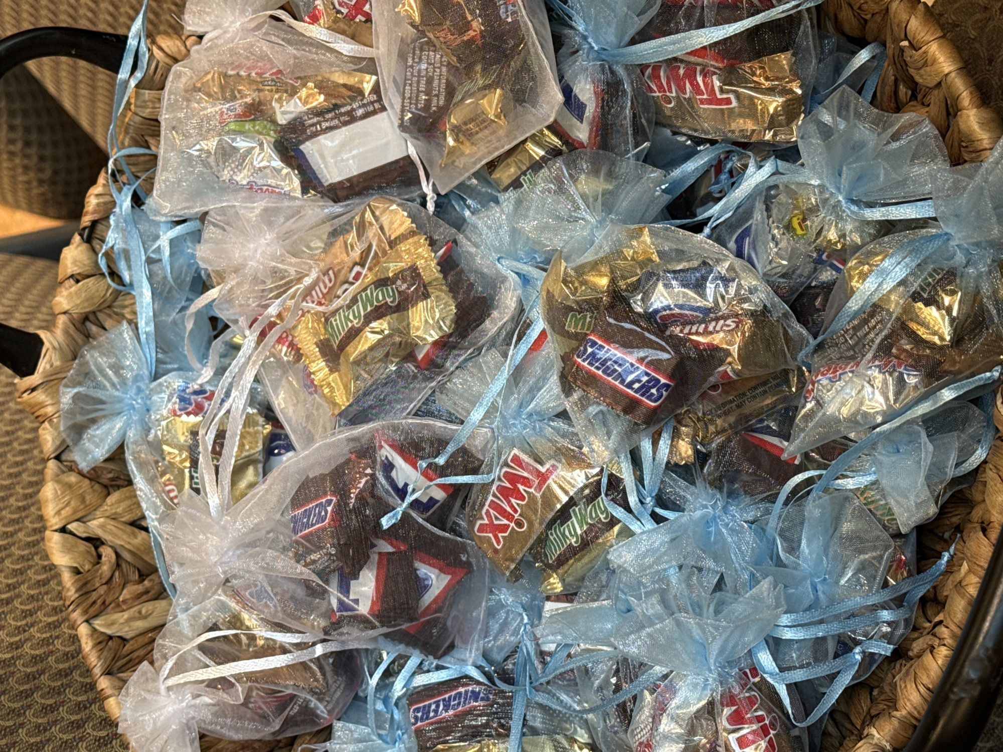 The image shows a close-up, top-down view of a large, brown wicker basket filled with numerous small, individually wrapped chocolate candies. Each candy is enclosed in a translucent organza bag, most of which are a light blue color and tied with matching ribbons. A few clear or white organza bags are also visible.
The basket appears to be brimming with these small favors or treats. Visible chocolate candy brands include "Snickers," "Twix," and "Milky Way," suggesting a selection of miniature chocolate bars from Mars, Incorporated. The candy wrappers are clearly visible through the sheer material of the bags.
Visible text on some of the wrappers includes nutritional information and allergen warnings such as "CONTAINS PEANUTS, MILK, AND SOY," "MAY CONTAIN TREE NUTS," and "MAY CONTAIN PEANUTS," and "WHEAT." Contact information like "Call 1-800-551-0702" and "Mars Inc." along with a product code "1380166 BL" are also discernible on some wrappers. The setting appears to be indoors, possibly on a surface with a patterned carpet or rug visible in the background, near what looks like a black metal handle of the basket. The scene suggests prepared gifts or treats for an event or gathering.