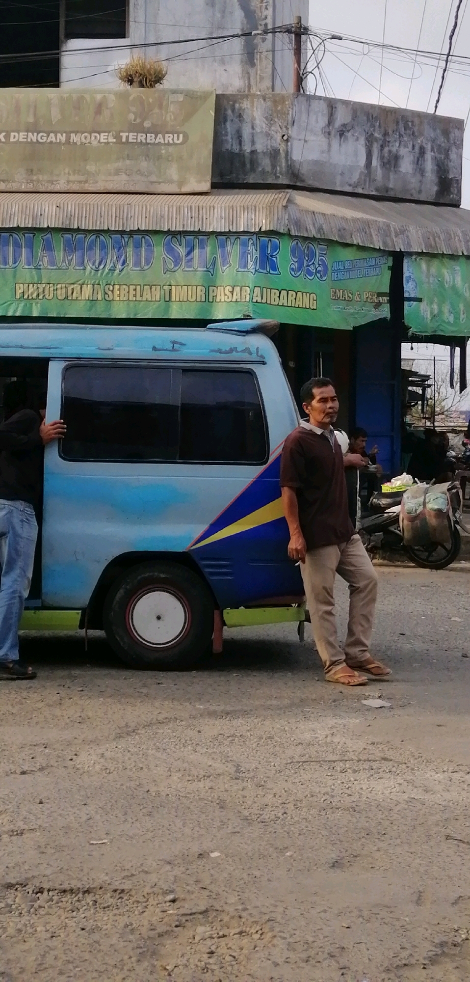 In Ajibarang, Indonesia, an outdoor scene unfolds with a light blue van parked on a dirt road. The van, decorated with a blue, yellow, and white diagonal stripe on its side, has its passenger door open. A person wearing blue jeans stands in the doorway, their left hand on the doorframe. To the right of the van, a man in a dark brown short-sleeved shirt and light brown pants leans against the vehicle. He is holding a cigarette in his right hand, which is near his mouth. Behind the man, a motorcycle with two large bags strapped to its rear is visible. Above the van, a banner with the text "DIAMOND SILVER 925" is prominently displayed, along with smaller text reading "PINTU UTAMA SEBELAH TIMUR PASAR AJIBARANG" and further details in Indonesian. The background includes buildings with grey, textured walls and overhead wires. The ground is a mix of dirt and gravel.