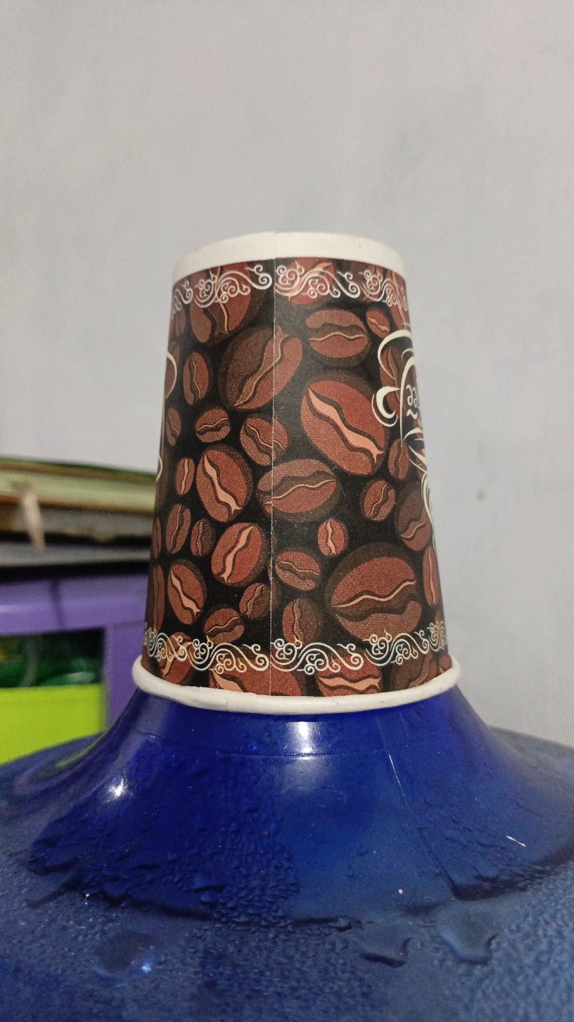 A close-up, low-angle shot shows a disposable coffee cup placed upside down on the blue cap of a water jug. The cup features a pattern of coffee beans and decorative borders. The water jug appears to be a standard dispenser bottle, with condensation visible on its surface. In the background, a blurred shelf with books and other items can be seen, along with a plain, light-colored wall. The scene is likely indoors, possibly in a home or office setting in Trihanggo, Indonesia.
