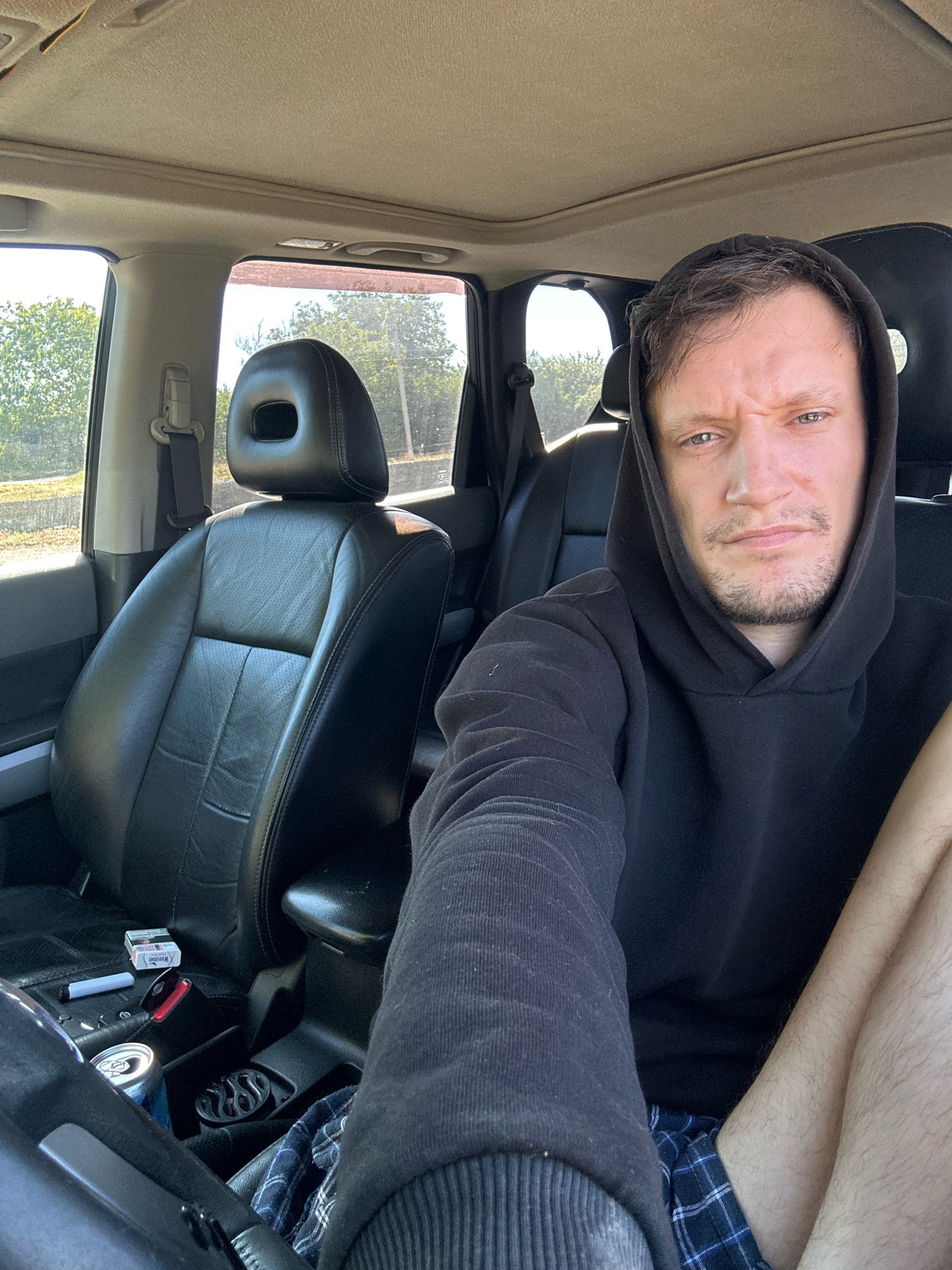 The image shows a man in the driver's seat of a car, possibly a Nissan X-Trail based on the interior. He is wearing a black hoodie and plaid shorts. On the car's center console are a pack of Winston cigarettes, a vape pen, a can of soda, and a car air freshener. The car's interior appears to be a light beige color for the headliner and darker for the seats and dashboard. The man is looking directly at the camera with a serious expression. Outside the car, trees and foliage are visible, suggesting an outdoor location, potentially in a rural area or on the outskirts of a town. There is no specific information in the image that places it in Makiv, Ukraine.