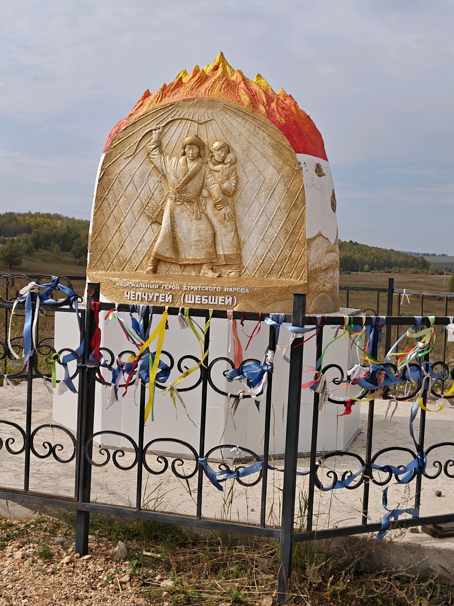 A golden monument depicting a man holding a bow and arrow and a child stands in front of a white cylindrical base. The monument is crowned with red and yellow jagged peaks, resembling flames or mountains. The inscription below the figures reads "NATIONAL HERO OF THE BURYAT PEOPLE CHEPCHUGEY (SHEBSHEI)". A black metal fence with decorative scrollwork surrounds the monument. Colorful ribbons are tied to the fence, fluttering in the breeze. The background shows a rural landscape with rolling hills covered in trees, a field, and a cloudy sky. This monument is located in the village of Bayandai, Russia.