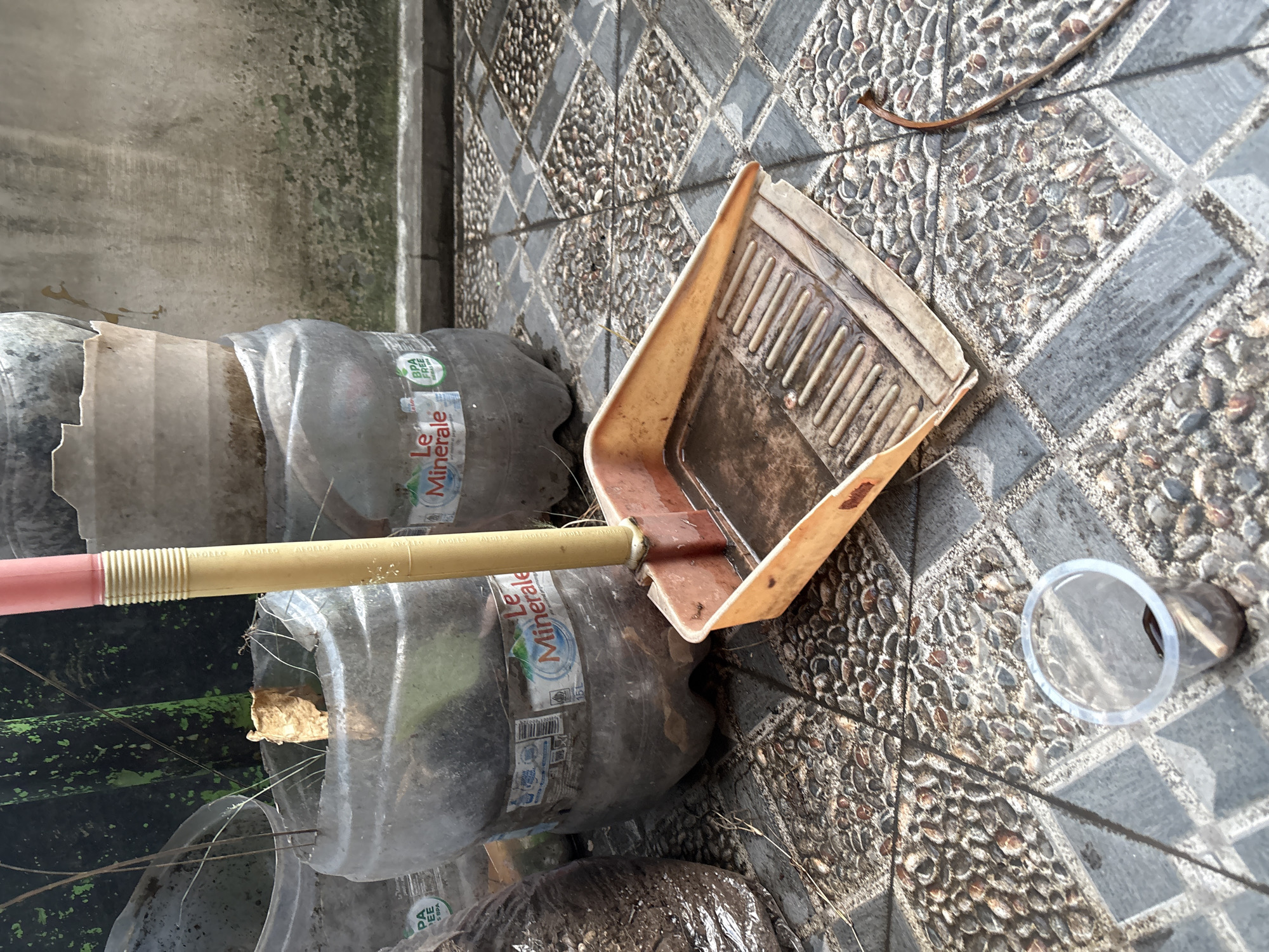 An outdoor scene in Palimanan, Indonesia features a close-up of gardening equipment and repurposed plastic bottles. A dustpan with a yellow handle lies on a patterned tiled floor. The dustpan contains water and debris. Several large, clear plastic bottles, likely repurposed as planters, are positioned behind the dustpan. One bottle has a "Le Minerale" label. The environment appears to be a residential or garden area.