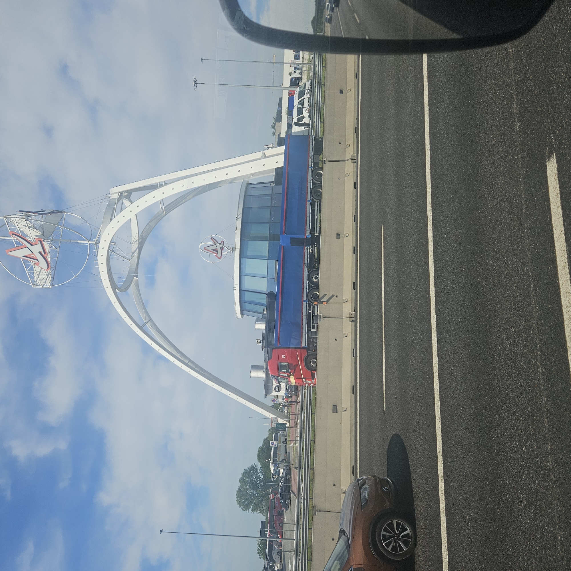 The image captures a view from a vehicle traveling on a highway. A large, white, arched structure is visible to the left of the highway, featuring a sign with a red "A" logo within a circle. This is the iconic entrance arch to the Alfa Romeo Museum, located in Lainate, Italy. Adjacent to the highway, there is a line of parked semi-trucks and other vehicles beyond a fence. The sky is partially cloudy. The presence of the highway lanes and other cars indicates active traffic flow.