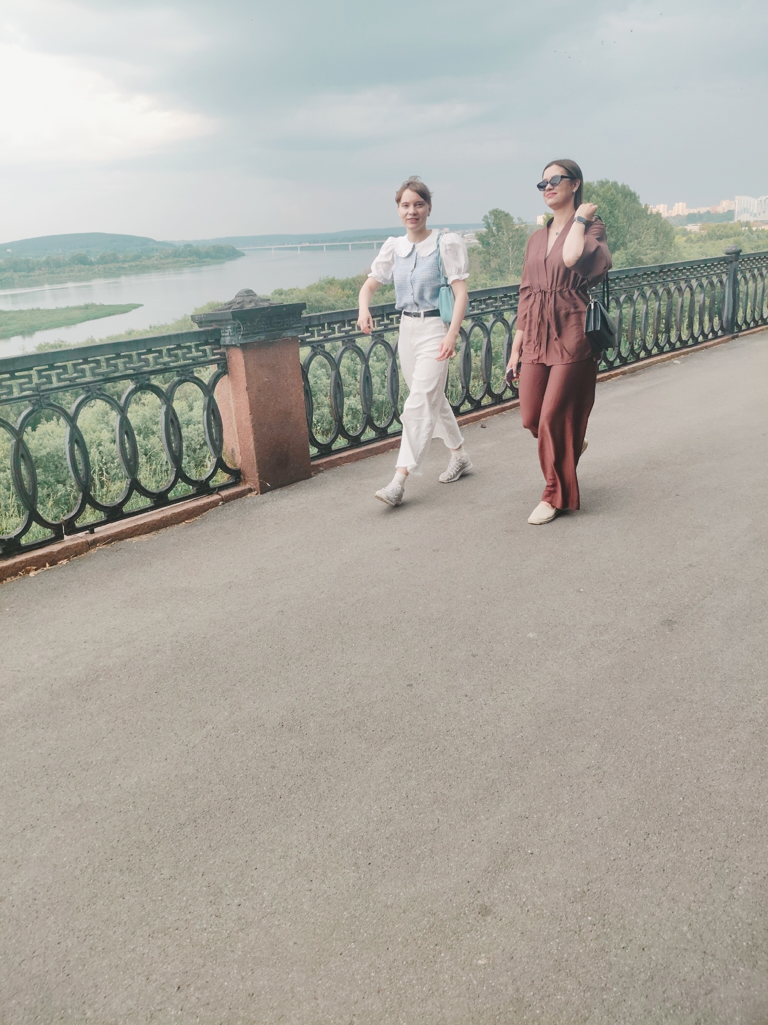 The image shows two young women walking along a paved pathway next to a decorative metal fence. The woman on the left wears a light blue and white patterned top with puffed sleeves and white pants, paired with white sneakers and socks. She carries a light blue crossbody bag. The woman on the right is dressed in a rust-colored jumpsuit and light beige flat shoes. She wears sunglasses and a watch on her left wrist, and carries a black shoulder bag.
They are walking on a paved area, possibly a promenade or sidewalk, with a view of a wide river and lush green hills in the background. A bridge can be seen spanning the river in the distance, and a city skyline with buildings is visible on the far bank. The weather appears overcast, with a cloudy sky. The scene evokes a leisurely outdoor activity, possibly a walk in a park or along a waterfront. The women appear relaxed as they stroll, with the woman on the right looking slightly off-camera to her right. The decorative fence features a repeating pattern of circles within arches, and the pathway is bordered by a stone wall. The overall setting suggests a scenic overlook in a city.
