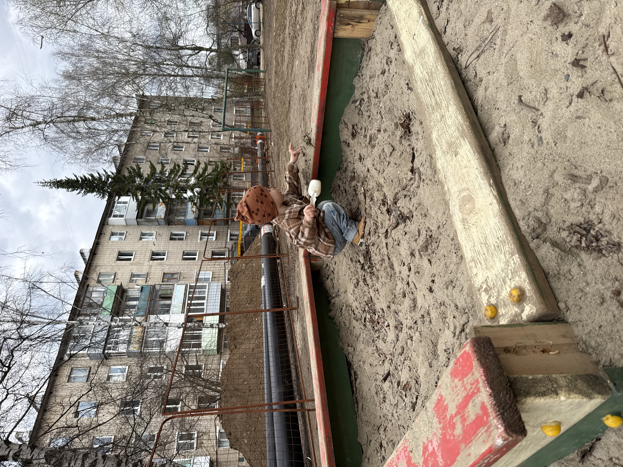 A high-angle, slightly Dutch-tilted photograph captures a young child playing in a sandbox in what appears to be an urban setting in Yaroslavl', Russia, under an overcast sky. The child, wearing a brown patterned hat, a plaid shirt, and blue jeans, is squatting in the sandy area, holding a white scoop or shovel in one hand and pointing upwards with the other.
The sandbox is defined by wooden planks, some of which are painted red and green, with yellow caps or details attached. The sand is light brown and appears somewhat coarse, mixed with fallen leaves and debris. To the left of the sandbox, beyond a metal fence, a trench reveals large black pipes, possibly for district heating or water, suggesting ongoing construction or utility work.
In the background, a large, multi-story apartment building with numerous windows and several enclosed balconies dominates the scene. Its facade is a light, somewhat weathered brick or concrete. Bare-branched trees, indicative of late winter or early spring, frame the building and reach into the cloudy sky. A tall, slender conifer stands prominently in front of the building. To the far left, another large, bare-branched tree is visible. No discernible text is visible in the image.