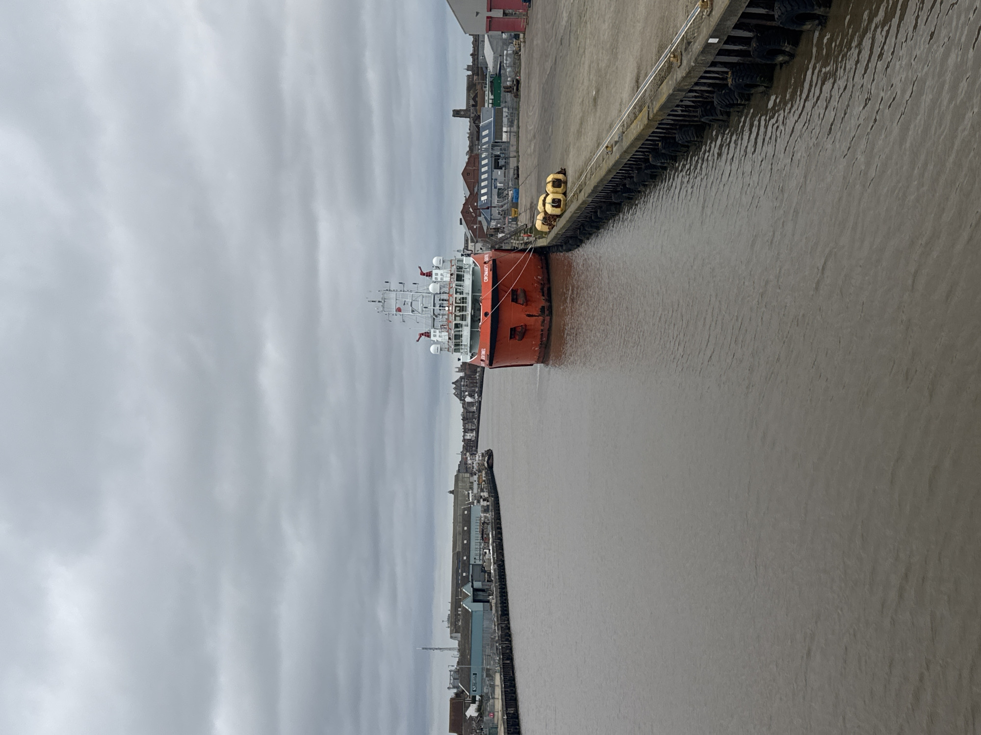 The image shows a cloudy day in Gorleston-on-Sea, United Kingdom. A large orange ship is docked along a concrete pier. Tires are attached to the pier to act as bumpers. Buildings are visible in the background on both sides of the water. The water is murky.