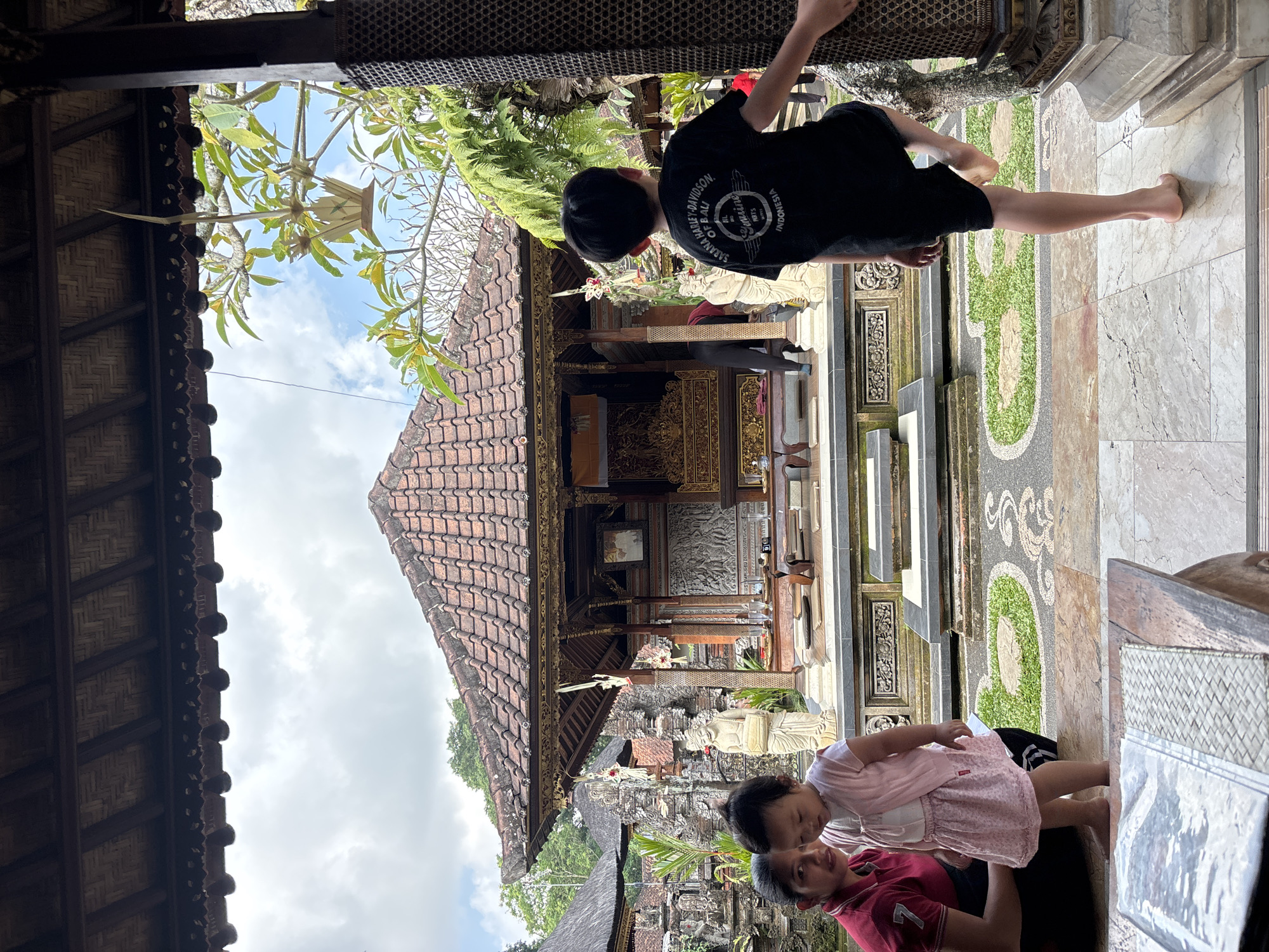 In Ubud District, Indonesia, a group of people are seen in what appears to be a traditional Balinese temple complex. The foreground features a young boy in a black t-shirt and shorts, facing away from the camera, with his right leg raised as if balancing. To his left, a woman in a red shirt and a young girl in a pink dress are partially visible.
The middle ground of the image showcases a meticulously designed temple structure with a tiered, terracotta-tiled roof and ornate wooden carvings. Statues and intricate stonework are visible around the temple, suggesting religious or cultural significance. Lush green foliage surrounds the buildings, hinting at a tropical environment. A pathway with patterned stone and grass leads up to the temple.
The background reveals more of the temple complex, with other structures and trees against a cloudy sky. The overall scene conveys a sense of cultural heritage and a serene atmosphere.
