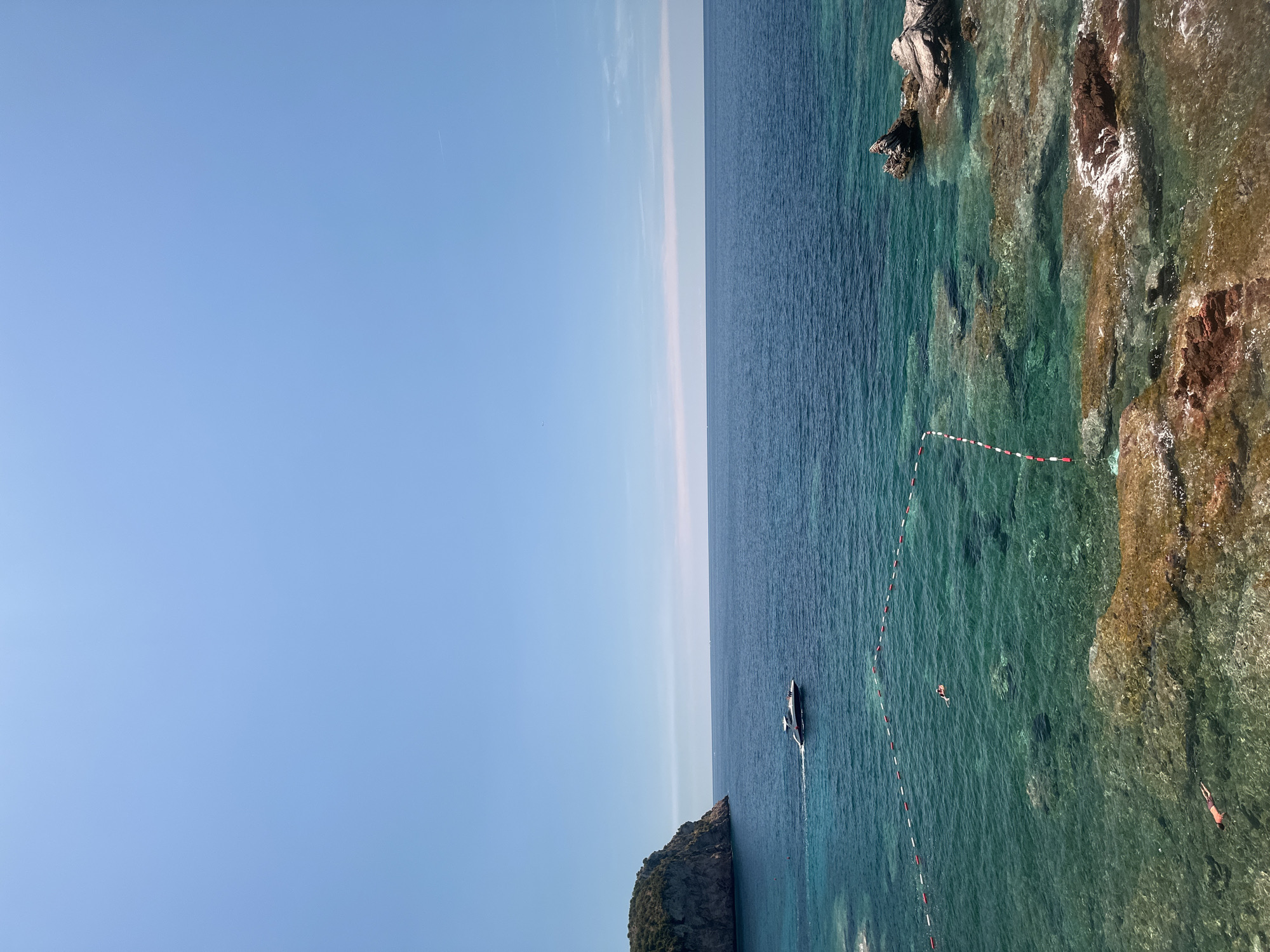 This image captures a scenic view of the Adriatic Sea near Sveti Stefan, Montenegro. The upper portion of the frame is dominated by a clear, pale blue sky, with a faint contrail visible in the distance on the right. The horizon line is sharp, separating the sky from the deep blue sea.
In the mid-ground, a white motor yacht is cruising across the water, leaving a subtle wake. To the left, a rugged, green-covered island juts out into the sea, its steep cliffs contrasting with the calm water.
The foreground reveals the shallow, crystal-clear turquoise waters close to the shore. The seabed is visible, showing a mix of sandy areas and rocky formations with reddish-brown hues. Buoys with alternating red and white markers delineate a swimming area in the water. Two individuals are visible swimming in this marked area. Another person is partially visible at the bottom left, near the shore, also appearing to be in the water. The overall impression is a tranquil and picturesque coastal scene on a clear, sunny day.