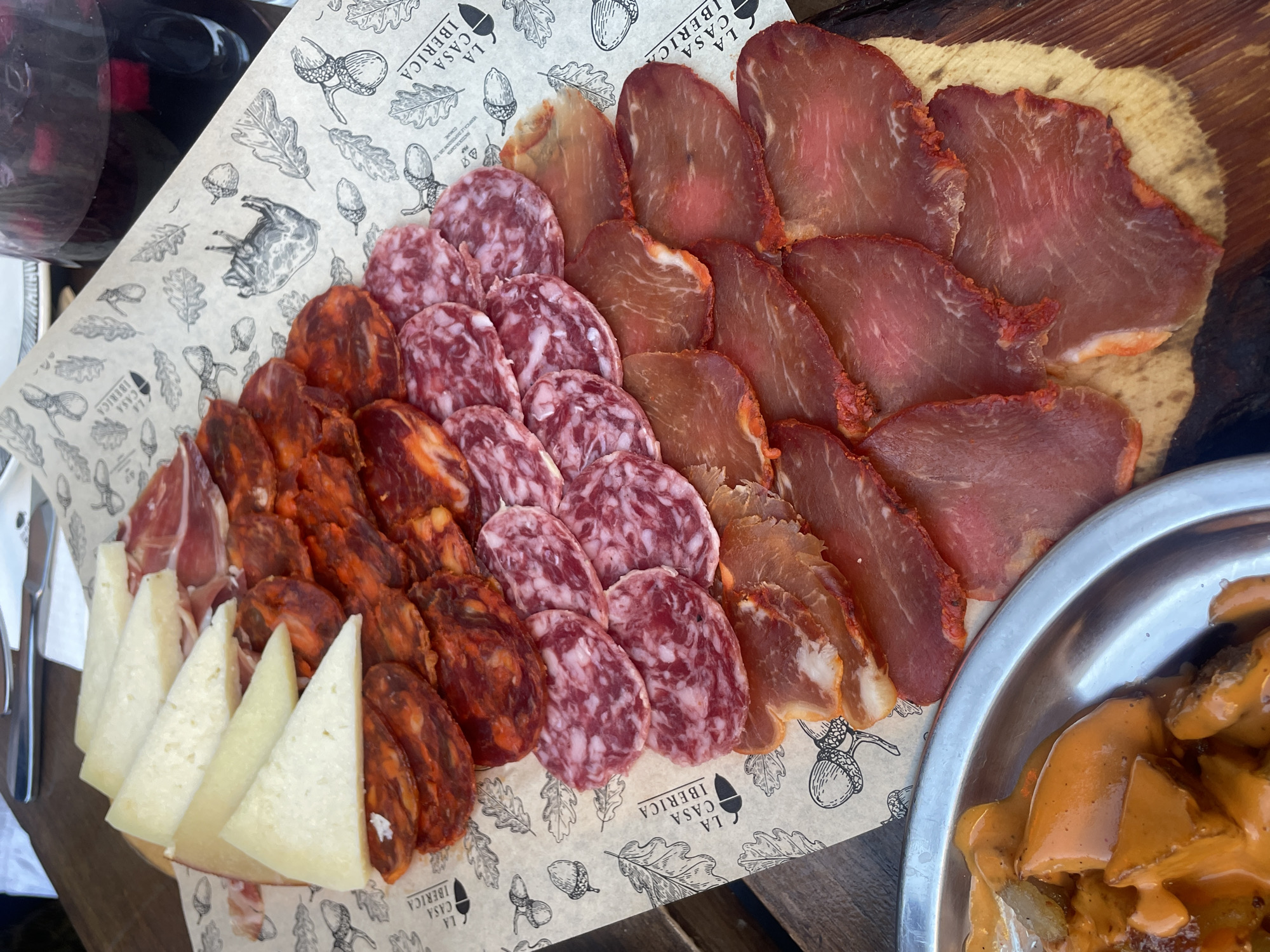 A wooden serving board is topped with a sheet of decorative paper patterned with oak leaves, acorns, and the text "LA CASA IBERICA." The board displays a selection of Spanish meats and cheese. Arranged in a semi-circular pattern are wedges of hard cheese, followed by slices of dark red chorizo, pink and white salami, lighter-colored cured meat, and finally, slices of deep red lomo. A silver tray containing food covered in an orange sauce sits beside the board. Cutlery is visible to the left, and a dark bottle is in the upper left corner. The scene appears to be set outdoors, possibly in a restaurant or at a picnic, in Milan, Italy.