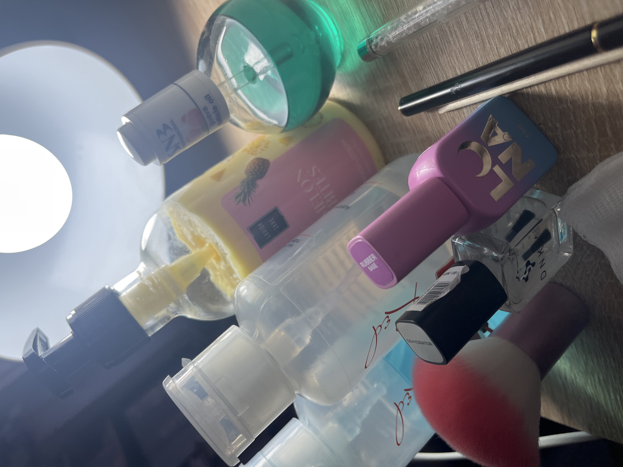 The image shows an assortment of nail care products arranged on a light-colored surface, possibly a tabletop. The products include bottles of cuticle oil, a pineapple-scented lotion, a pink-lidded nail polish with the inscription "RUBBER BASE," a nail dehydrator, a container with the brand "LG NA," and a makeup brush with pink bristles. The bottles are of varying sizes and shapes, with some labels visible. The products appear to be in a scattered, disorganized arrangement. The lighting is strong, possibly from a nearby light source causing highlights on the bottles. There is no indication of a specific location in Pernik, Bulgaria.