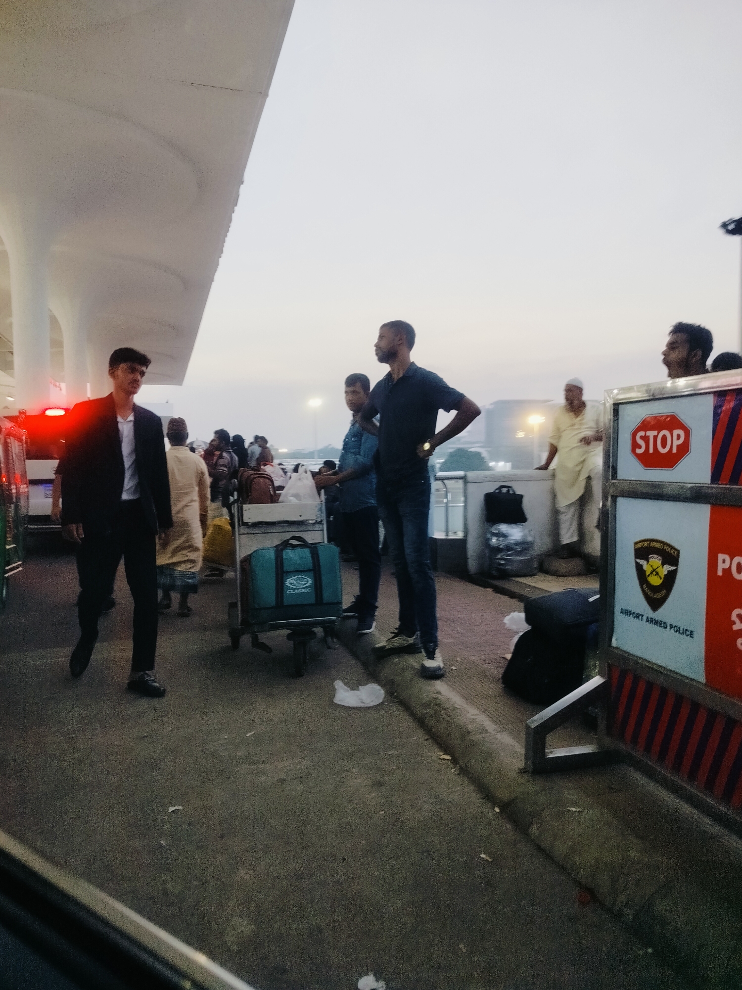 This image depicts an outdoor scene at Hazrat Shahjalal International Airport in Dhaka, Bangladesh. Several individuals are present, some with luggage. In the foreground, a man in a dark suit and white shirt walks towards the viewer. To his right, a luggage cart is visible with a teal-colored duffel bag and plastic bags. Further back, several other people are seen, some carrying bags and others standing. A man in a dark polo shirt and jeans stands with his hands on his hips, looking to the left. To the right of this man, a white barrier with a "STOP" sign and an "AIRPORT ARMED POLICE" emblem is present. The sky is overcast, suggesting it is either early morning or late afternoon. The environment appears to be a paved walkway or road, likely part of the airport's arrival or departure area. Red brake lights of a vehicle are visible on the far left.