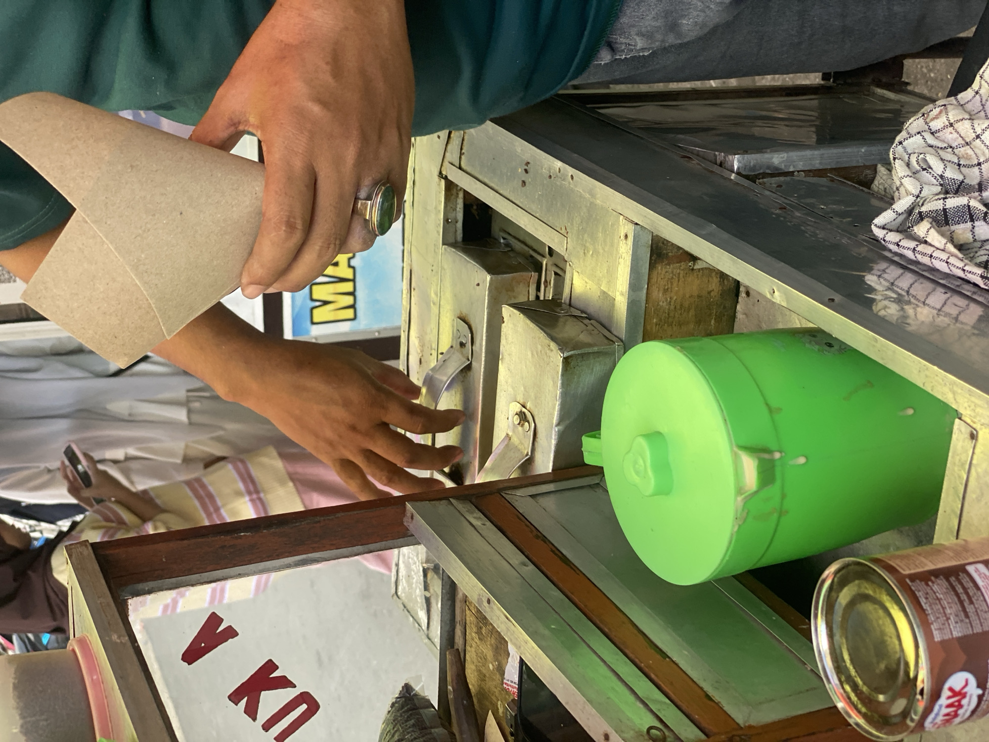 In the city of Banjarbaru, Indonesia, a close-up shot captures a street vendor at work. A person, whose hands and torso are visible, is holding a cone made of brown paper, appearing to prepare a treat. The hands are adorned with a ring featuring a green stone. The vendor is positioned behind a mobile food cart, which is constructed from metal and wood. A prominent bright green plastic container with a lid sits on the cart, with some white liquid dripping from its rim, suggesting it holds a beverage or ingredient. Adjacent to it is a metal can, partially obscured, with a label that includes red lettering. The cart's surface is metallic, with some compartments visible. In the background, out of focus, are glimpses of other individuals and what appears to be a striped shirt and headscarf, hinting at a bustling street scene. The overall impression is one of a typical Indonesian street food vendor operating in a local setting.
