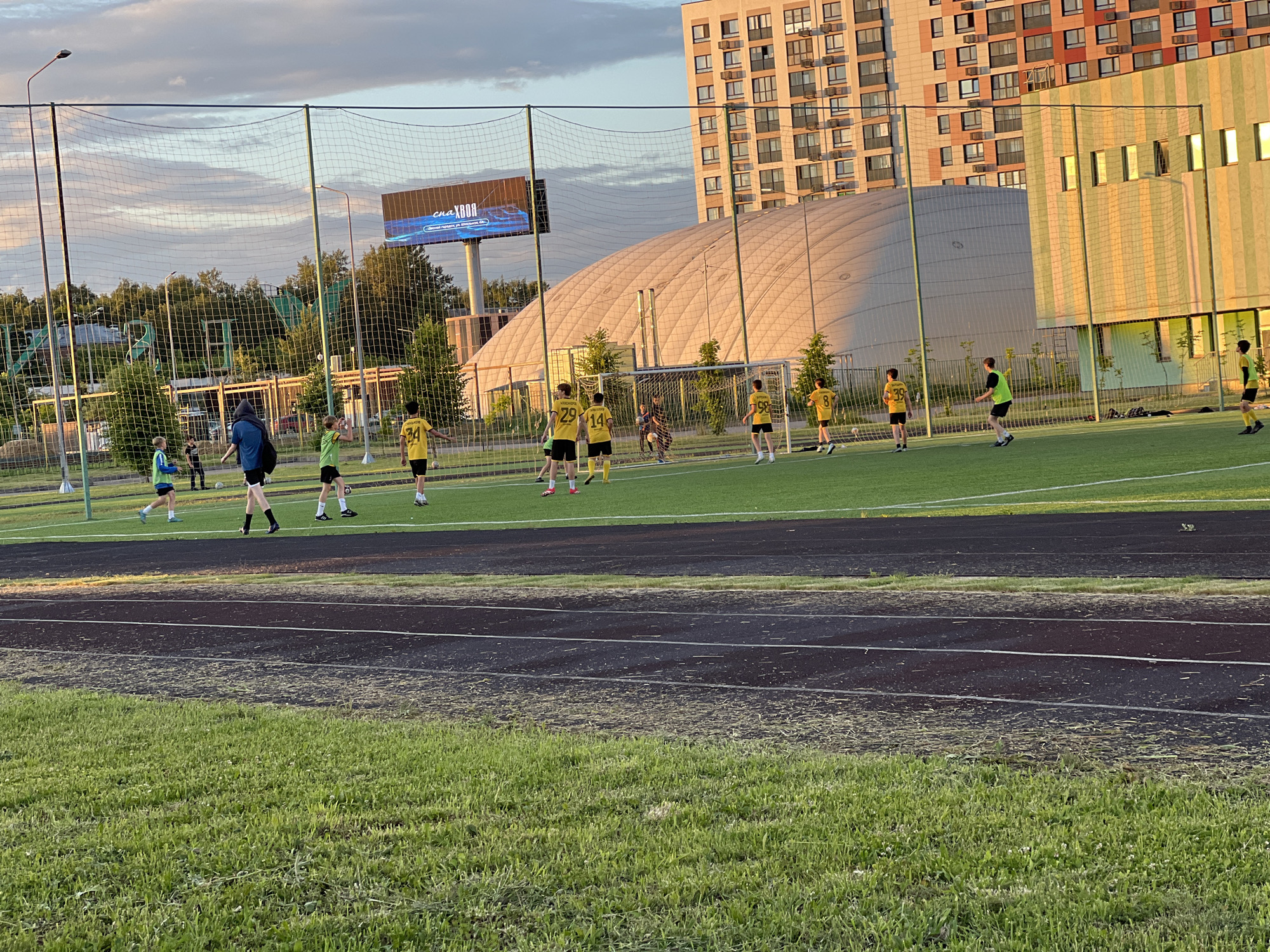 The image shows a group of young people playing soccer on an outdoor turf field. The field is enclosed by a net, and there are stadium lights surrounding it. In the background, there are tall apartment buildings and a large, rounded building with a striped facade. The sky is cloudy with hints of sunset colors. Some of the players are wearing yellow jerseys with numbers, while others are in green or blue. One person is wearing a black hooded jacket. The game appears to be in progress, with players actively engaged in running and interacting with the ball. The overall scene depicts a recreational activity taking place in an urban environment.