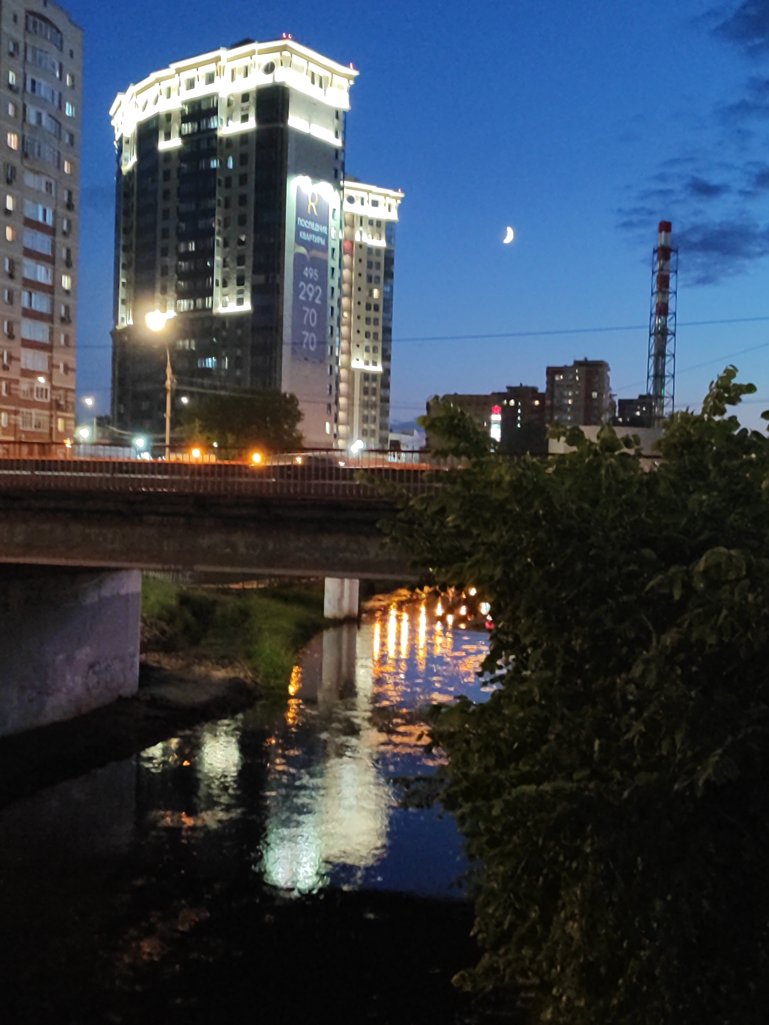 In the city of Unknown, Unknown, the nighttime scene is dominated by the reflections of illuminated buildings in a dark body of water. Two modern, tall buildings, brightly lit with white and yellow lights, stand prominently in the background. One building displays a large banner with Cyrillic text and numbers: "R ПОСЛЕДНИЕ КВАРТИРЫ 495 292 70 70". To the right, a tall industrial structure with red and white bands is visible. A crescent moon hangs in the deep blue sky. A bridge crosses the foreground, with streetlights and building lights reflecting on the water below, creating streaks of orange and white light. Lush green foliage occupies the lower right portion of the image, obscuring some of the scene. The overall atmosphere is one of urban nighttime ambiance.