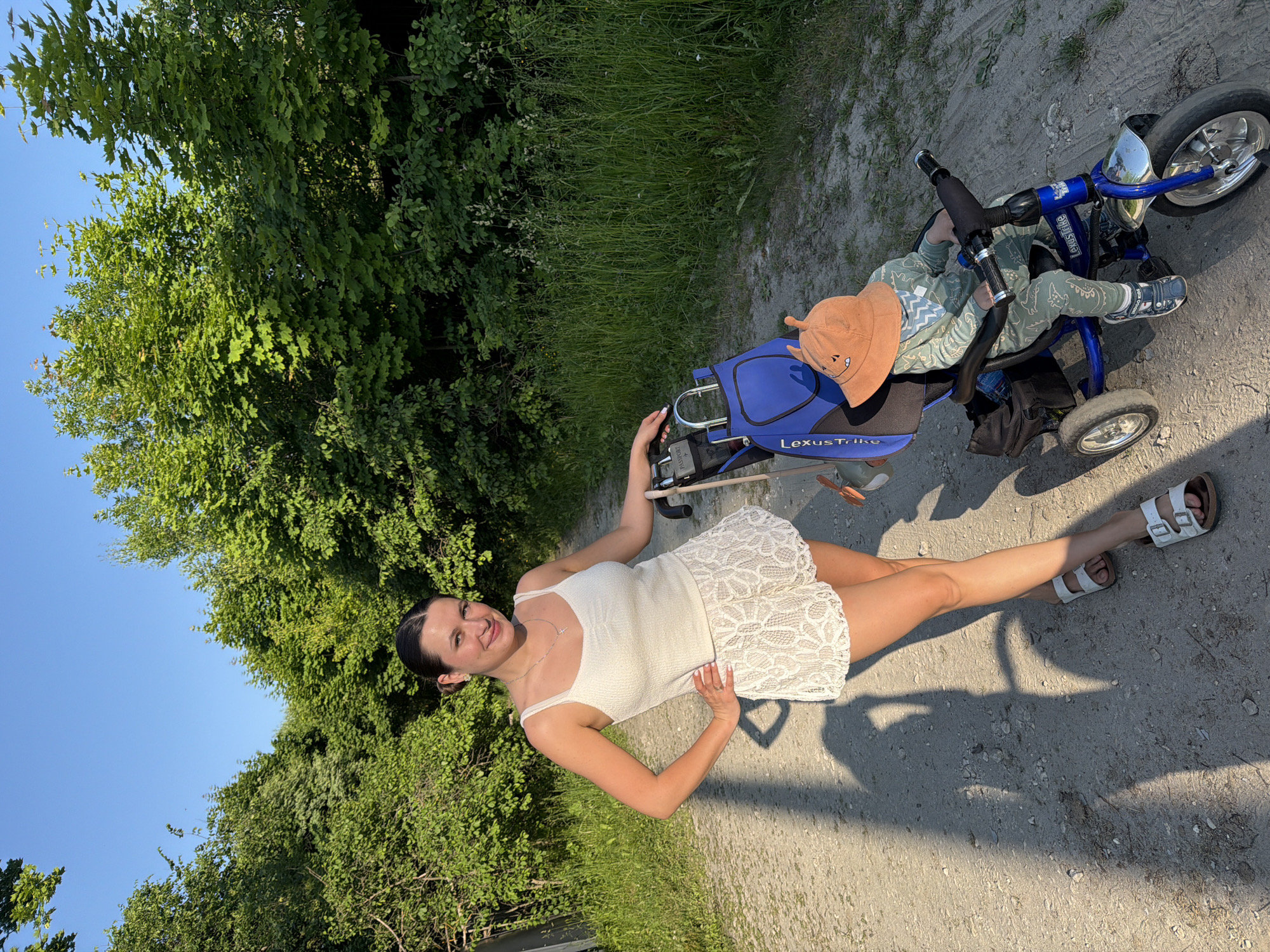 A woman and a child are outdoors on a dirt path with trees and grass on either side. The woman, wearing a white tank top and white lace shorts, stands with her hand on her hip, smiling at the camera. Next to her, a child sits on a blue tricycle wearing a green dinosaur-patterned outfit and an orange bucket hat. A blue bag is attached to the back of the tricycle. The sun is casting long shadows on the path, suggesting it is late afternoon or early morning. The location is identified as Krasny Mayak, Russia.