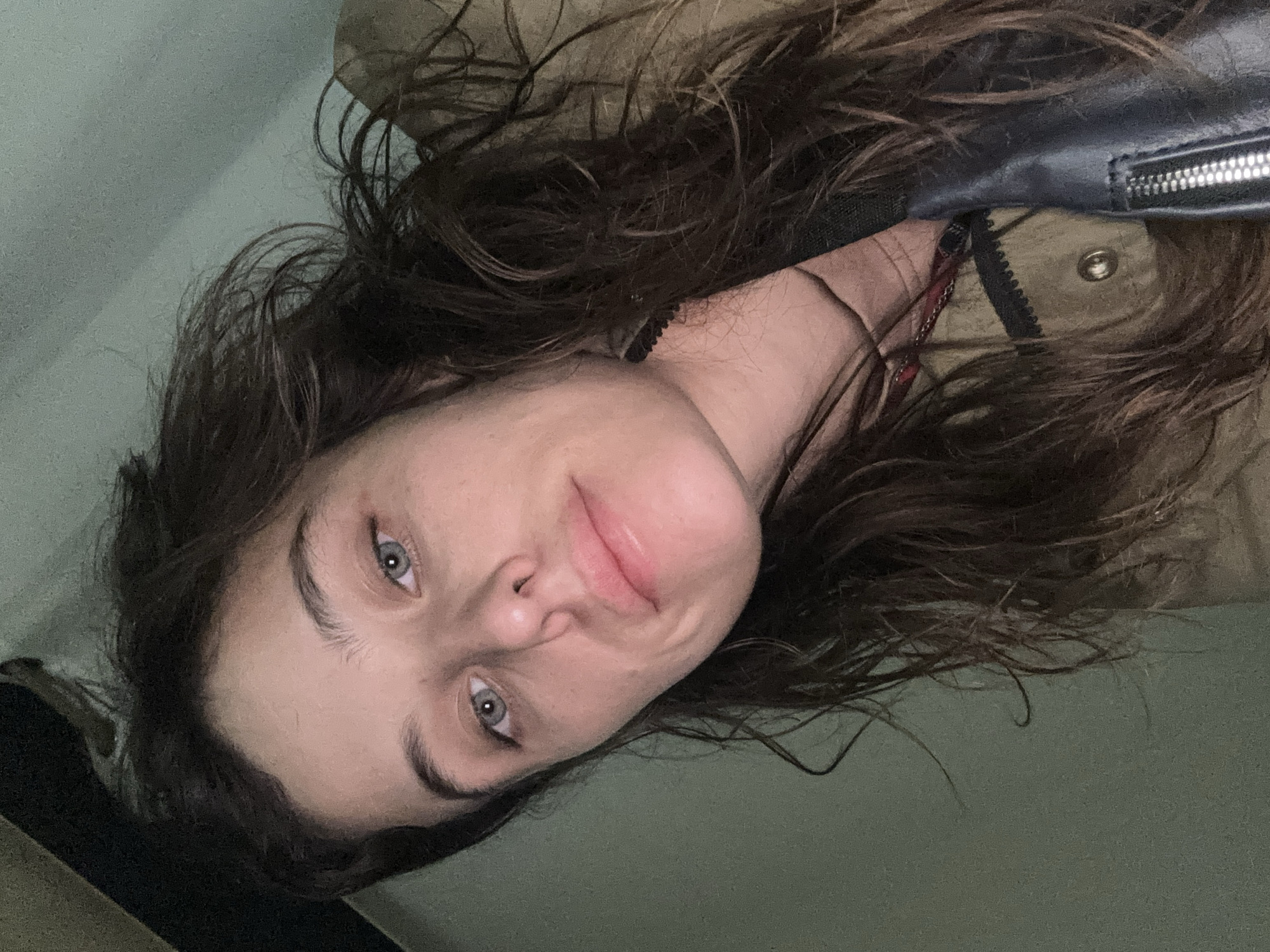 A close-up, downward-facing selfie of a young woman with dark, wavy hair and light blue eyes is captured. She is wearing a tan jacket with black trim and a black strap across her chest. Her lips are slightly parted, and she has a neutral expression. The background is a soft, muted green, suggesting an indoor environment. There is no indication of the specific location in Seogwipo-si, South Korea, in the image.