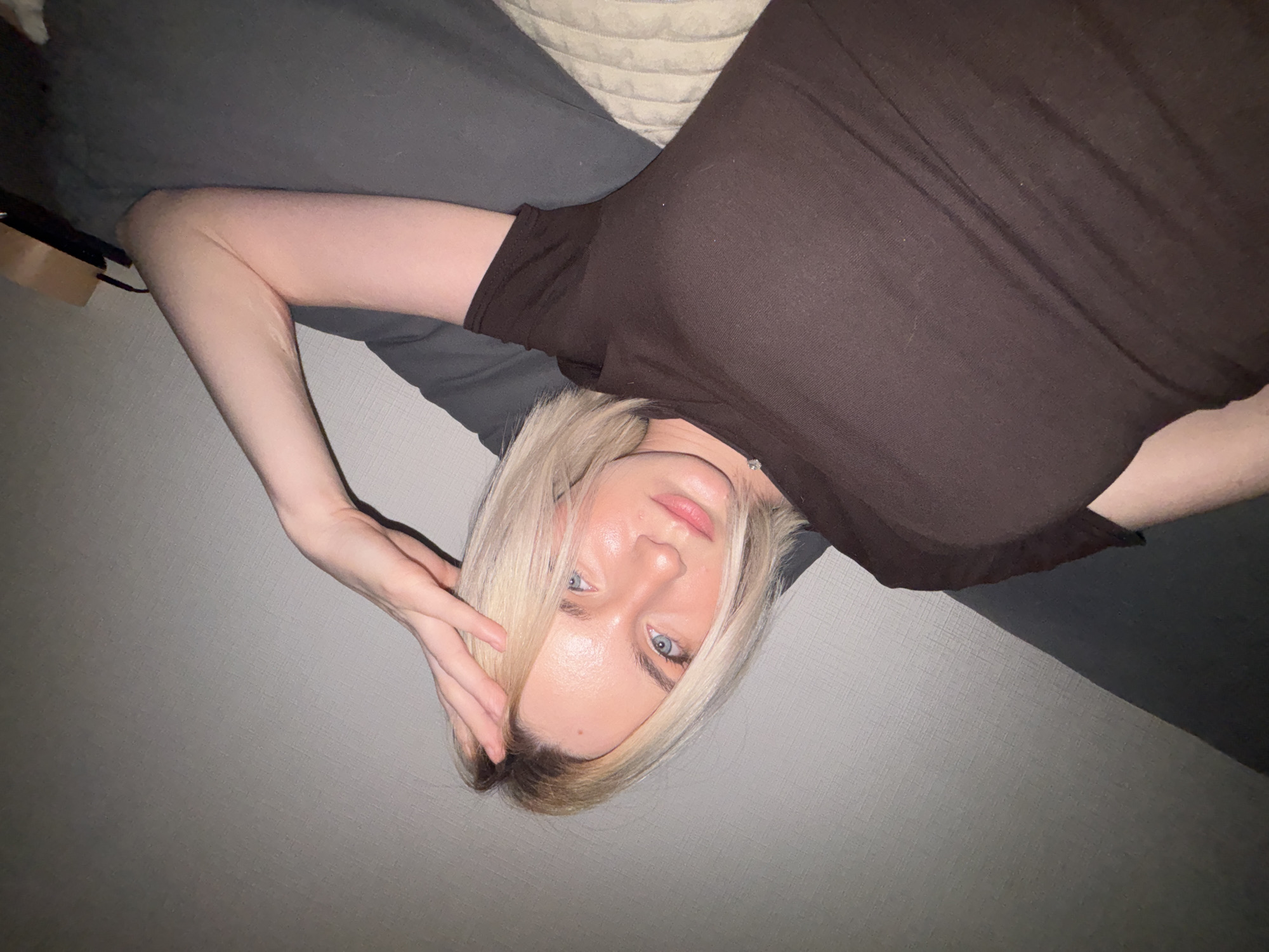 A fair-skinned woman with blonde hair and blue eyes lies on a gray surface, possibly a bed or couch. She wears a brown t-shirt and is positioned at an angle to the camera. Her left arm is raised, with her hand near her forehead. The background is a gray textured wall. There is no indication of the location being Vsevolozhsk, Russia.