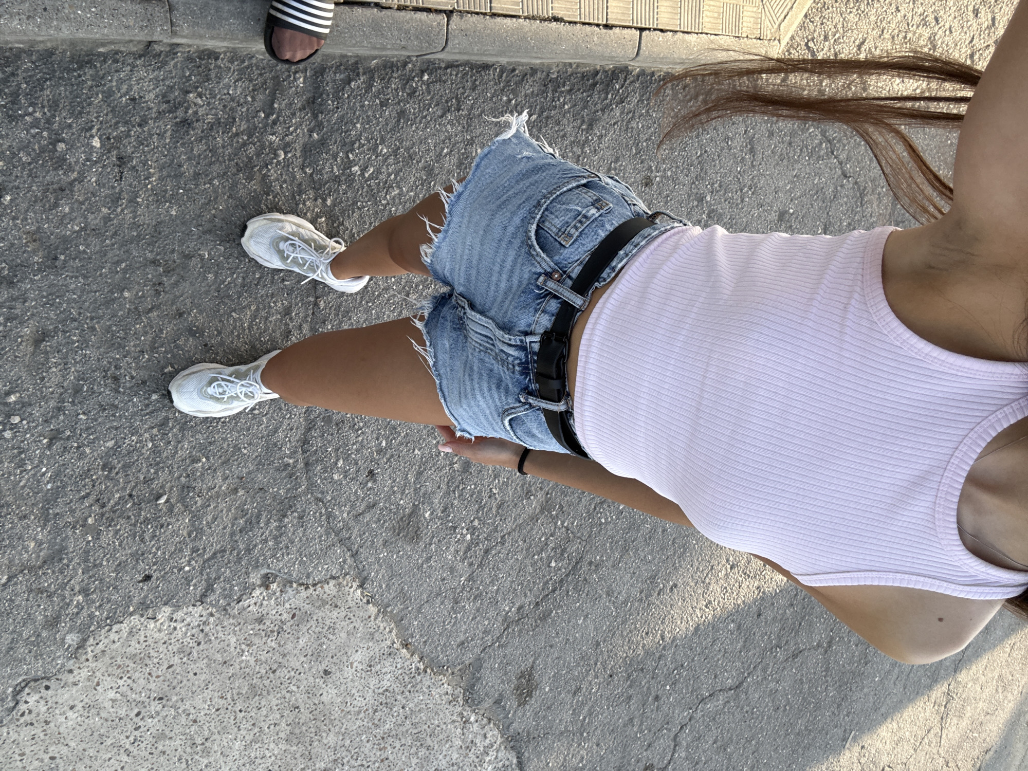 A person, viewed from a high angle, stands on a paved surface in Cimișlia, Moldova. The person is wearing a light pink ribbed tank top, light blue denim shorts with frayed hems, a black belt, and white athletic shoes. Their long brown hair extends downwards. The ground is a mix of asphalt and concrete, with some cracks visible. A shadow falls across the ground from the upper left. Another person's feet are visible in the lower right corner, wearing black and white striped slides and dark socks.