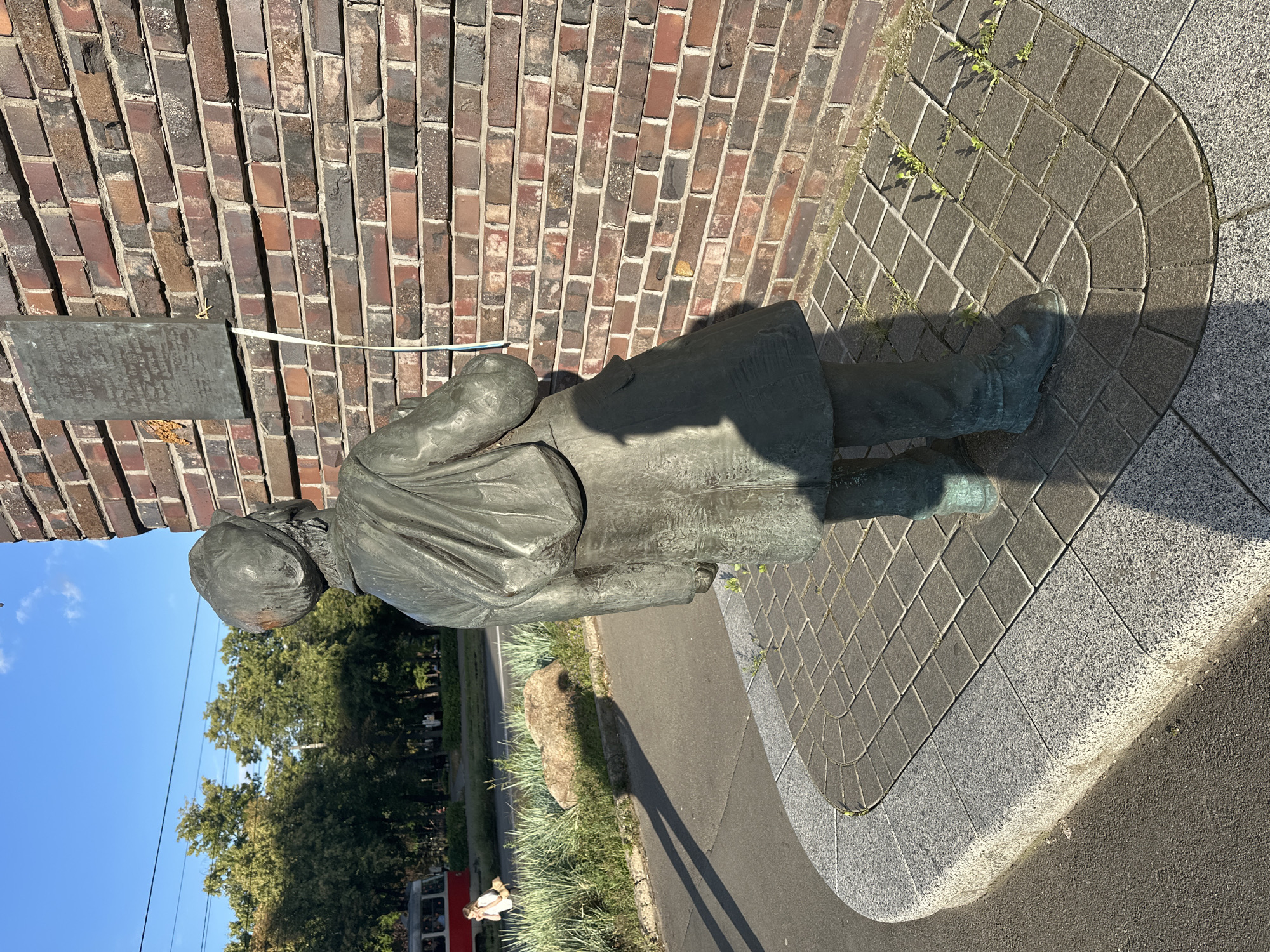 A bronze statue of a person is prominently displayed outdoors, leaning against a textured brick wall. The statue depicts a figure wearing a long coat and hat, with its body positioned at a slight angle. A rectangular metal plaque is attached to the brick wall near the statue. The ground around the statue is paved with diamond-shaped stones, bordered by a curved granite edge. Sunlight casts a shadow from the statue onto the pavement. In the background, lush green trees and a clear blue sky are visible. A red tram or train with windows can be seen in the lower left portion of the image, suggesting an urban or suburban setting. The scene appears to be illuminated by daylight, likely in the morning or afternoon, given the bright sunlight and clear sky. No specific emotion is discernible from the statue. There is no visible text in the image that can be clearly read.