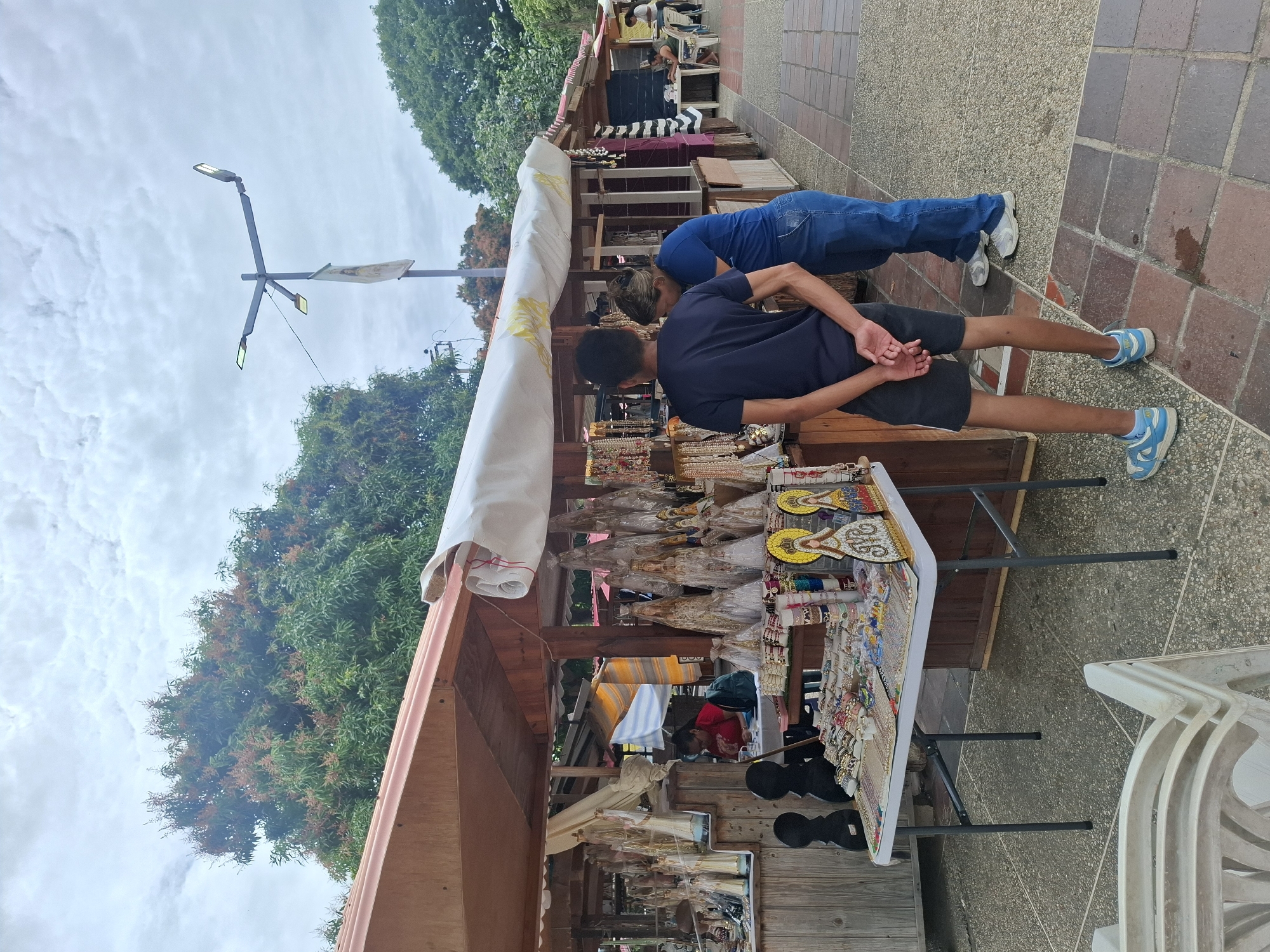 The image captures an outdoor market scene in El Valle del Espíritu Santo, Venezuela, under an overcast daytime sky. A wooden market stall, partially covered by a white fabric canopy, dominates the left side.
On a table within the stall, a diverse array of souvenirs and crafts is displayed. These include numerous small religious figures, possibly virgins or saints, many adorned in white and gold. Colorful beaded jewelry, such as necklaces and bracelets, is meticulously arranged, alongside various decorative trinkets. Some larger, yellow, fan-shaped items are also visible, one clearly displaying the name "MARIA," and other figures feature partially legible text, possibly "VENEZUELA."
Two individuals are seen from behind, browsing the items at the stall. The person closer to the foreground is a male wearing a dark blue t-shirt, dark shorts, and light blue sneakers, with his hands clasped behind his back. Behind him, another individual in a blue long-sleeved shirt, blue jeans, and white sneakers stands nearby, also seemingly engaged with the market. Further back within the stall's structure, other people are visible, including one person in a red shirt attending to merchandise and others seated on chairs.
The ground is a paved walkway constructed from reddish-brown and grey-brown interlocking bricks. In the background, dense green trees with some reddish blooms or foliage line the area. A modern lamppost with multiple light fixtures, possibly solar-powered, stands tall, with a small white banner or cloth tied to it. The sky is filled with soft, scattered clouds. The scene conveys a relaxed shopping atmosphere at a local craft market.