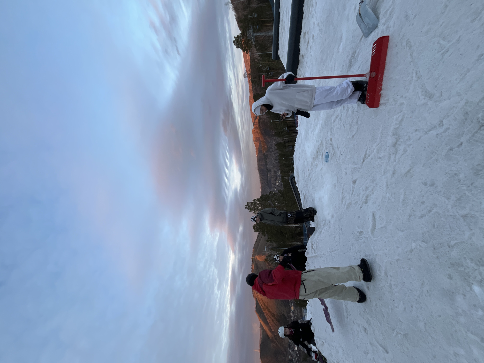 At a snow resort, a person in white attire is using a red snow removal tool. Several individuals are standing with skis and snowboards. The background displays a mountain range with an orange-tinted sky.