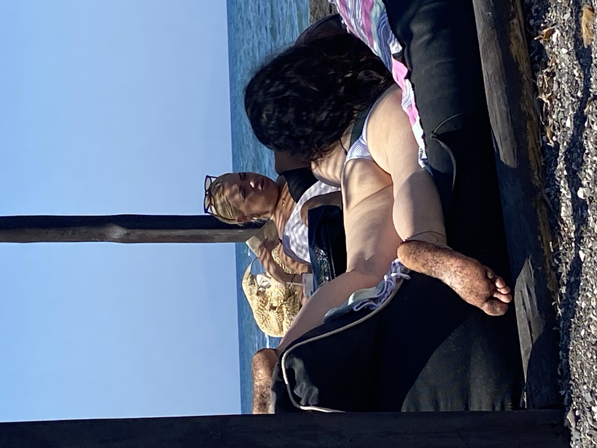 Two women are relaxing on a large black cushion on a beach. The woman in the foreground has her back to the camera, and her legs and feet are visible. Her feet are covered in sand. She is wearing a striped bikini bottom. The woman behind her is wearing a white top and a black bikini top, and has sunglasses on her head. The sea and sky are visible in the background. The scene appears to be outdoors on a sunny day.