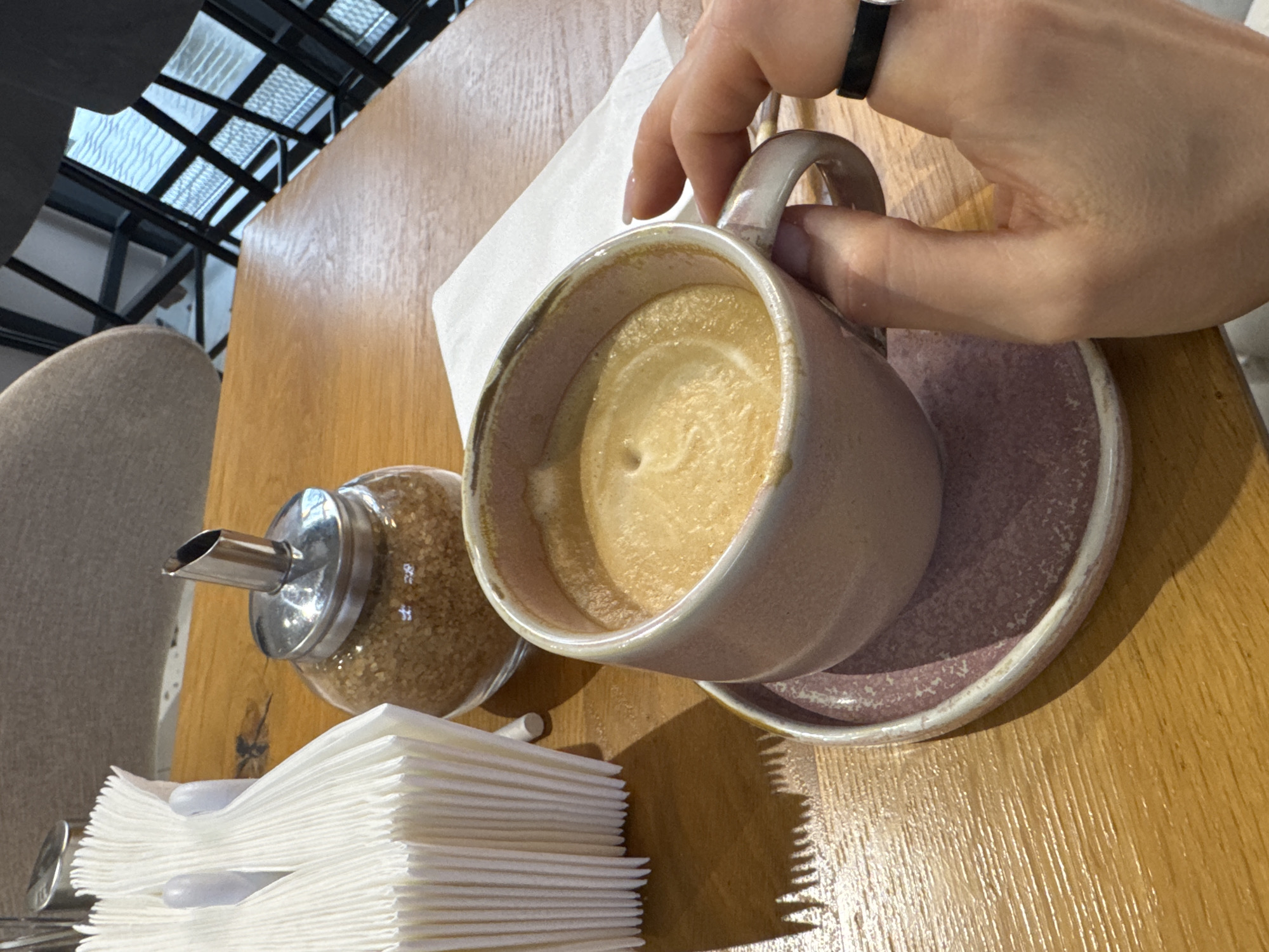 A coffee cup and saucer are on a wooden table next to a sugar dispenser and napkins. A hand with a black ring is holding the cup. The location is a cafe or restaurant in an unknown city.