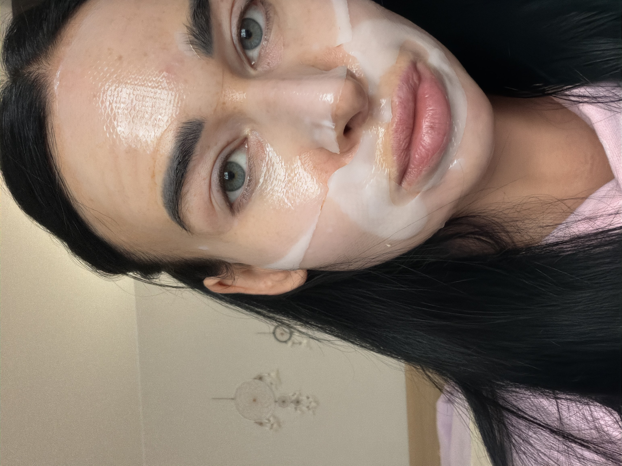A woman with dark hair is looking directly at the camera. She has a white sheet mask applied to her face in sections, leaving portions of her forehead, cheeks, and neck exposed. The background appears to be an interior setting with a ceiling fixture visible in the lower-left corner, suggesting a residential space. There is no information in the image to support a location in the city of Saint Petersburg, Russia.