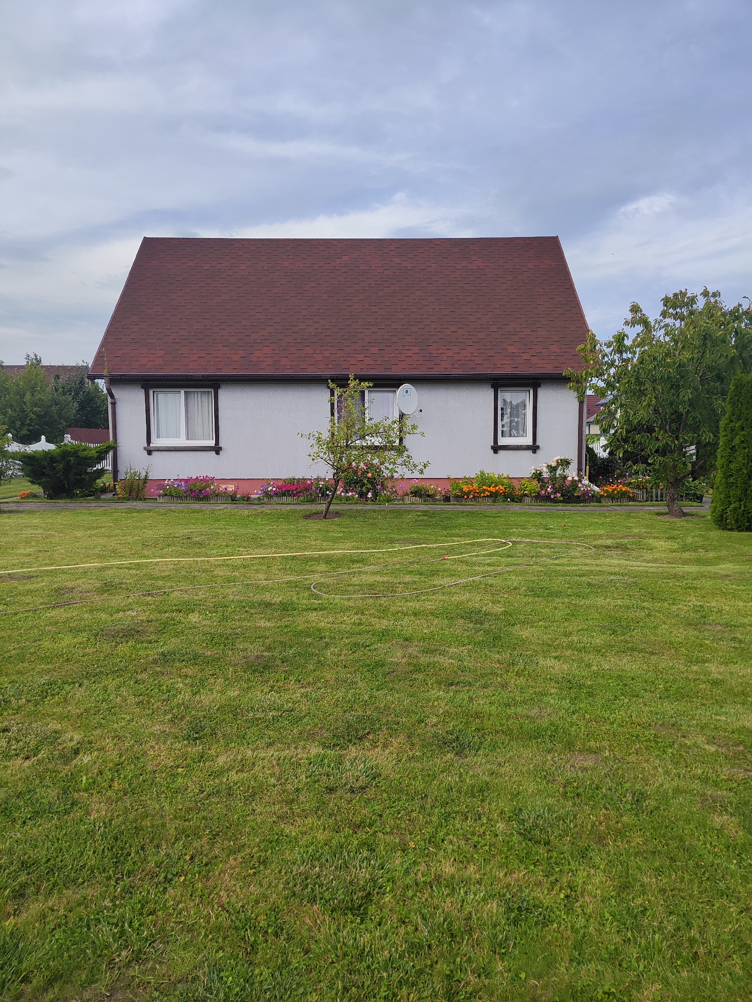 The image shows a single-story house with a red shingled roof, located in Gvardeysk, Russia. The house has light gray siding and two windows on the front facade, each with a white frame. In front of the house, there is a flower bed with various colorful flowers. A small tree with pink and orange blossoms is situated in front of the right window. The yard is covered in green grass, with a yellow hose visible on the lawn. The background features trees and a cloudy sky.