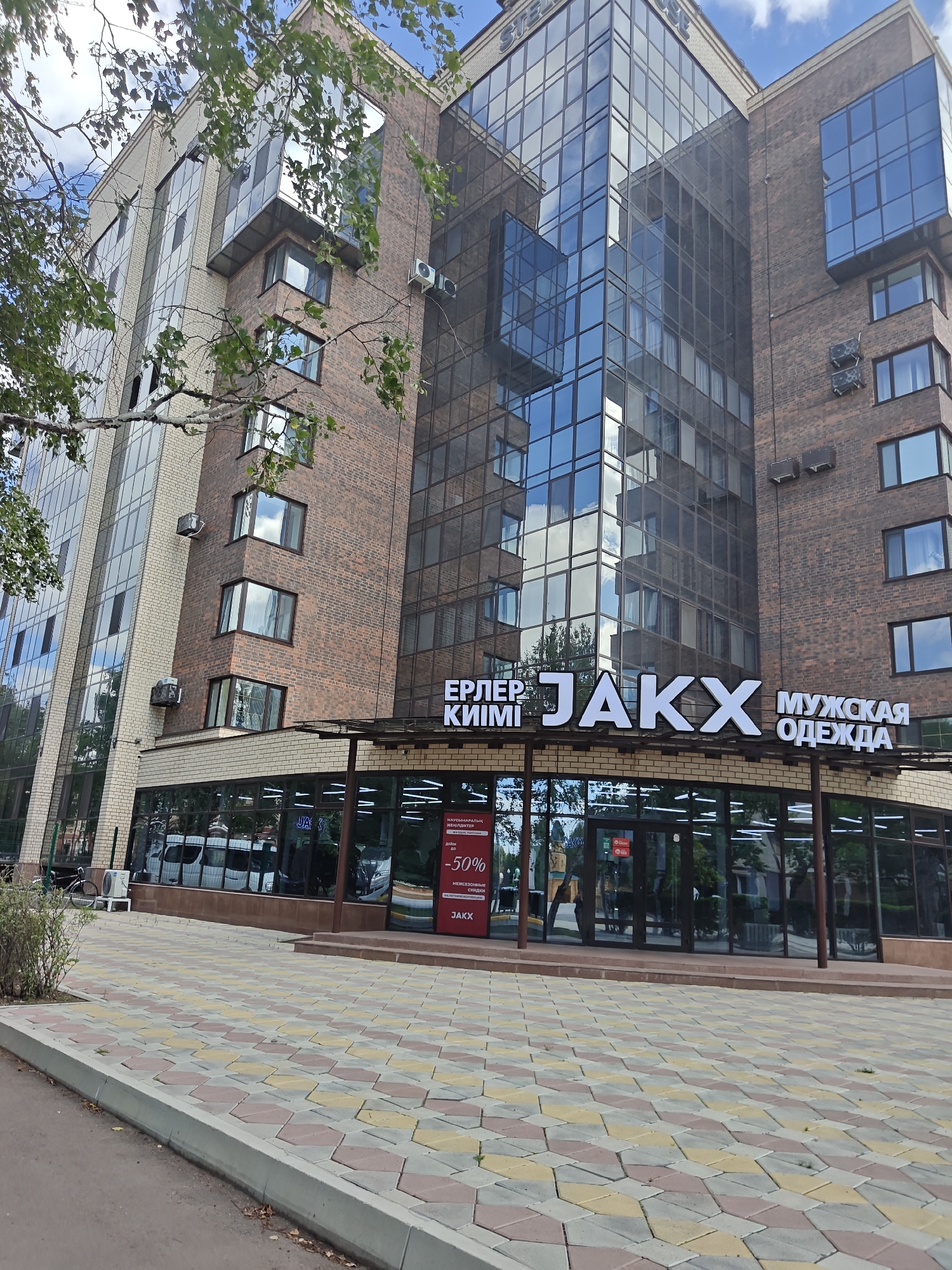 The image shows a modern, multi-story building with a glass facade and brick exterior, located in the city of Petropavl, Kazakhstan. The building appears to be a commercial establishment. A sign above the ground-floor entrance reads "ЕРЛЕР JАKX МУЖСКАЯ ОДЕЖДА," indicating it is likely a men's clothing store. The ground floor features a large glass storefront with reflections of trees and the sky. A sidewalk made of interlocking hexagonal paving stones in shades of pink, yellow, and gray runs in front of the building. To the left of the entrance, a bicycle is parked next to an air conditioning unit. The sky is partly cloudy. There are no people visible in the immediate vicinity of the storefront.