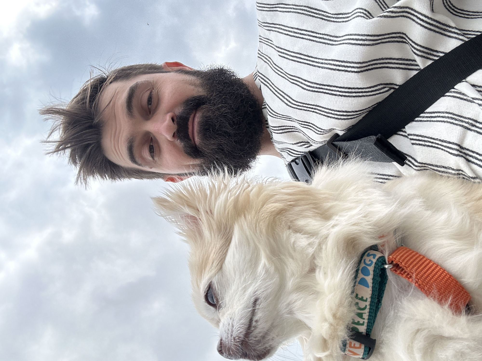 A person with a beard and messy hair is taking a selfie with a fluffy light-colored dog. The person is wearing a striped shirt and a black strap across their chest. The dog is wearing a collar that says "LOVE PEACE DOGS" and has an orange tag. The background is a cloudy sky. The image was taken in Khryashchevka, Russia.
