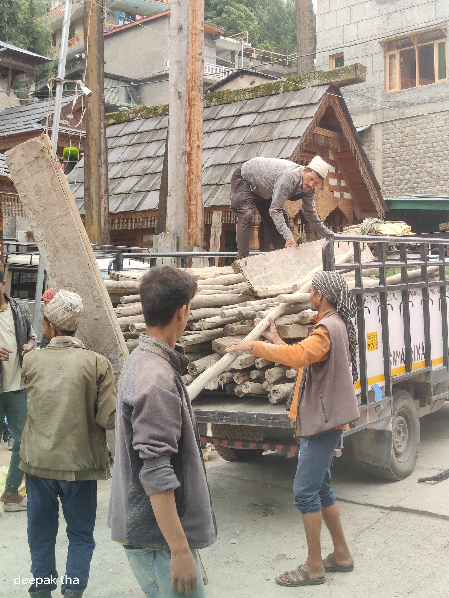 This image depicts a scene in Vashisht, India, likely a rural or mountainous area given the architecture. The foreground shows several men engaged in what appears to be manual labor, possibly loading or unloading materials from a truck.
In the center right, a man wearing a brown vest over an orange shirt and blue pants is holding a long, wooden log, with his gaze directed to the left. His head is covered with a checkered scarf. To his left, a man in a gray jacket and blue pants is turned to the right, with his back mostly to the camera, his right arm extended. In the left foreground, a man in a green jacket and blue pants is also facing away from the camera, with his left arm outstretched.
Behind these men, a truck is loaded with a large quantity of wooden logs. A man in a white cap and gray shirt is standing on top of the truck, bending down to handle some of the material. The truck itself has a registration plate visible, which appears to read "HP 58 A 8763". Various texts are printed on the side of the truck in white lettering on a yellow background.
The background reveals traditional Himachali architecture, characterized by wooden detailing and sloped, tiled roofs. Buildings are constructed with stone and wood, and the overall setting suggests a mountainous environment with vegetation visible on the slopes above. The ground is paved with dirt and gravel. The lighting suggests it is daytime.