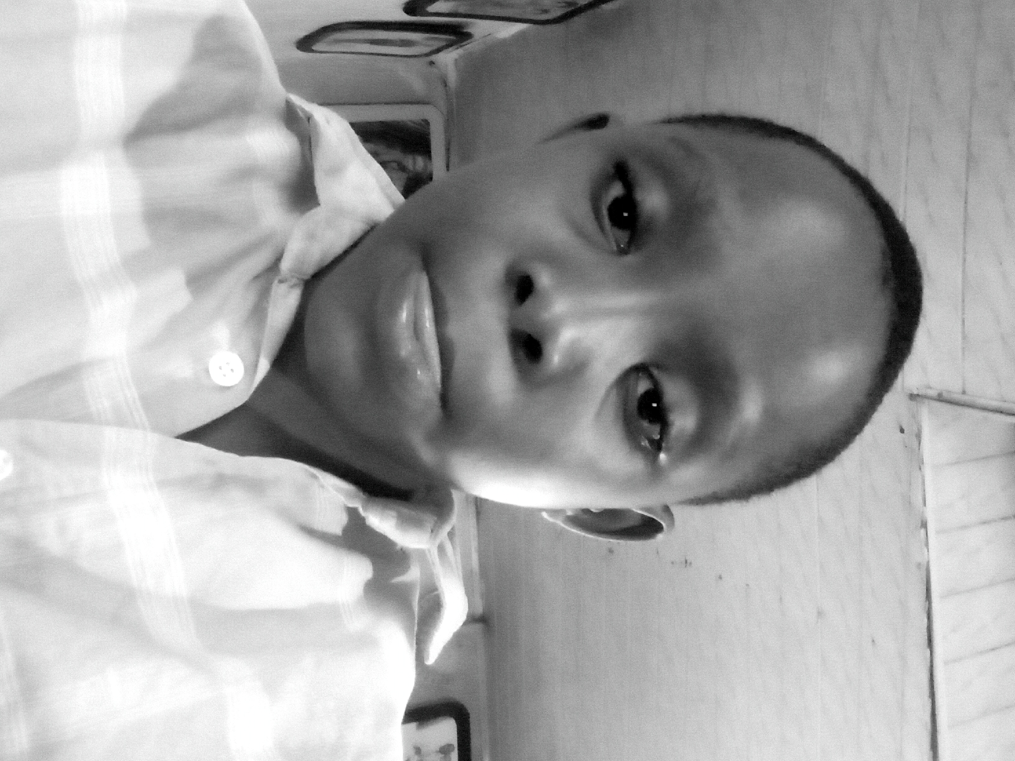 A close-up, black and white, medium shot selfie shows a young Black boy in a collared shirt. The boy's head is turned slightly to his right, and he is looking directly at the camera. His lips are slightly parted, and his eyes are open. His hair is short and dark. The shirt appears to be light-colored with a subtle striped pattern. The background is out of focus but appears to be an indoor setting with a wooden or paneled ceiling and walls. There are framed pictures visible on the wall to the left of the boy. The lighting is somewhat harsh, creating shadows on the boy's face.