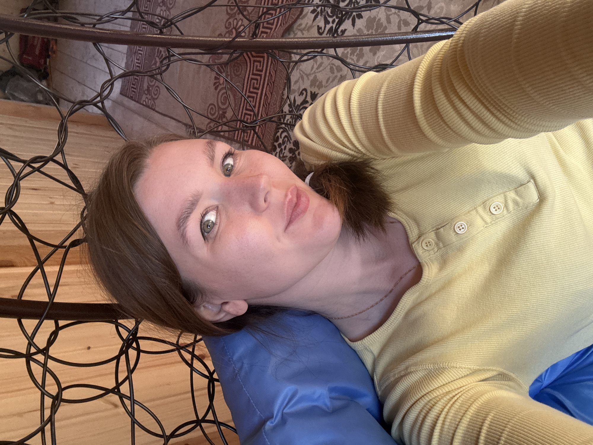 A woman with light skin and brown hair is lying down on a blue cushion. She is wearing a yellow, button-down shirt. The background includes a patterned rug, wooden walls, and a metal frame structure with circular designs. The photograph was taken indoors. The specific location, Italmas, Russia, cannot be determined from the visual information provided.