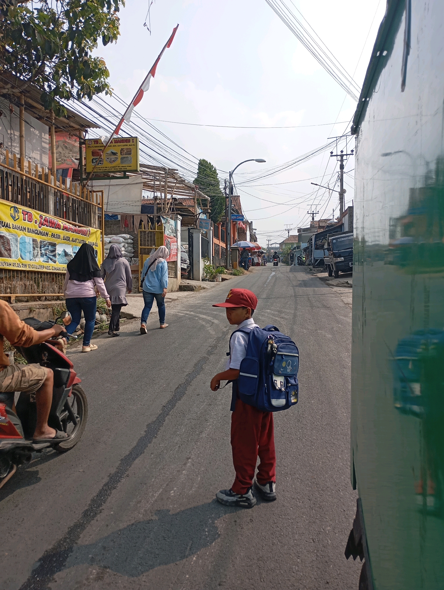 A street scene unfolds in Cimahi, Indonesia, under a bright, overcast sky. A young boy, dressed in a white school shirt and red pants with a red baseball cap, stands facing left, his back to the camera, carrying a large blue backpack adorned with space-themed patches. He is on the right side of the asphalt road. To his left, three women walk away from the camera, all wearing hijabs. The woman in the foreground wears a black hijab and a pink top with blue jeans. The middle woman wears a grey hijab and a grey tunic over black pants. The furthest woman wears a light blue hijab and a light blue shirt with blue jeans. A red scooter is parked on the left side of the road, with a person's legs visible sitting on it, wearing tan shorts and flip-flops. Various commercial buildings line the left side of the street, including one with a yellow banner displaying "TB. JAWA MANDIRI" and advertising building materials. Overhead, a dense network of electrical wires crisscrosses the sky, supported by poles. A large, dark green vehicle, possibly a truck, occupies the far right edge of the frame, its reflective surface showing a blurred image of the surroundings. In the distance, more buildings and parked vehicles suggest a bustling urban environment. A red and white Indonesian flag is partially visible on a pole near the buildings on the left.