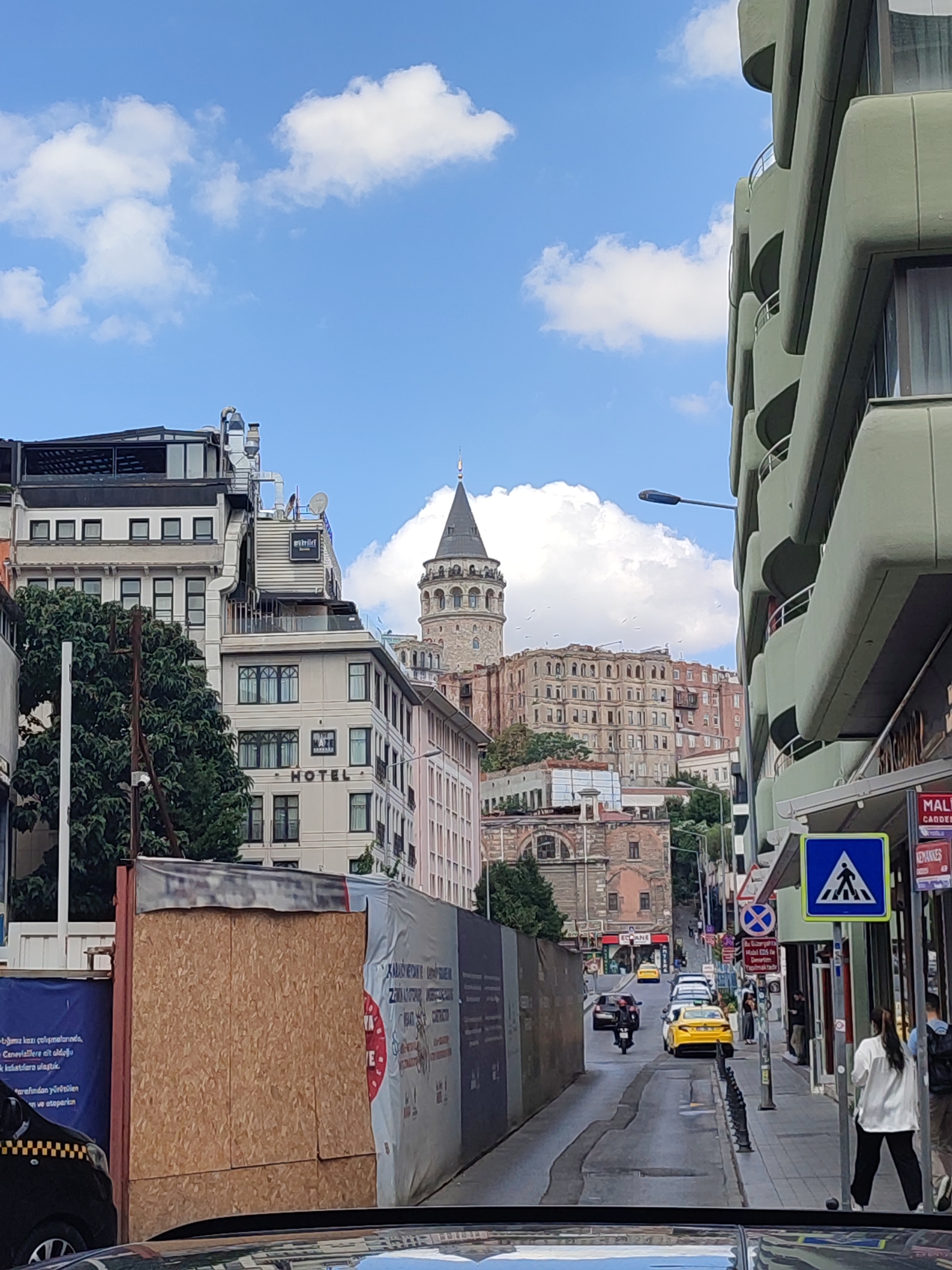 This image depicts a street scene in Beyoğlu, Turkey, dominated by urban architecture and a prominent historical tower. The Galata Tower, a medieval stone tower, stands in the background, its conical roof piercing a cloudy blue sky. In the foreground, a street slopes downhill, lined with buildings of varying architectural styles. To the right, a modern, curved building with green facade and balconies adds a contemporary element. On the left, older buildings with multiple windows and a sign reading "HOTEL" are visible. Construction barriers with plywood and sheeting are present on the left side of the street, suggesting ongoing development or renovation. Several vehicles, including a yellow taxi, a motorcycle, and other cars, are on the street, indicating moderate traffic. Pedestrians are also present, walking on the sidewalks. The overall scene captures a blend of historical and modern urban life in Beyoğlu.