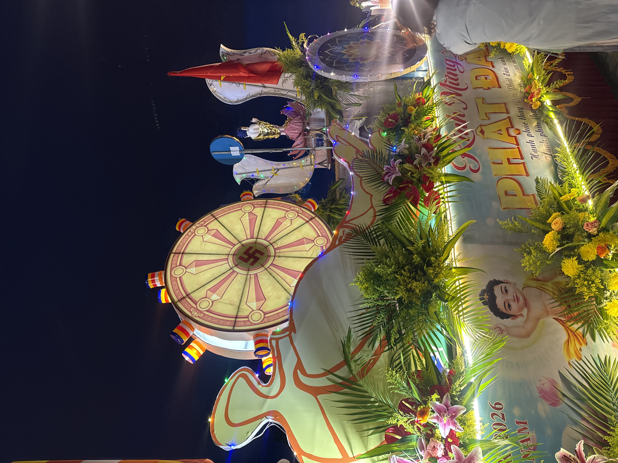 Captured at night in Da Lat, Vietnam, the image features an elaborate religious display. A large, illuminated wheel-like structure emblazoned with a symbol resembling a Swastika dominates the scene. Below the wheel are colorful, cylindrical lights. A poster with the words "PHẬT ĐẠ" is adorned with flowers and foliage. The poster features a depiction of a child and text in Vietnamese. The event seems to be a religious celebration based on the decorations and symbols present. A person partially obstructs the scene at the top right corner of the image.