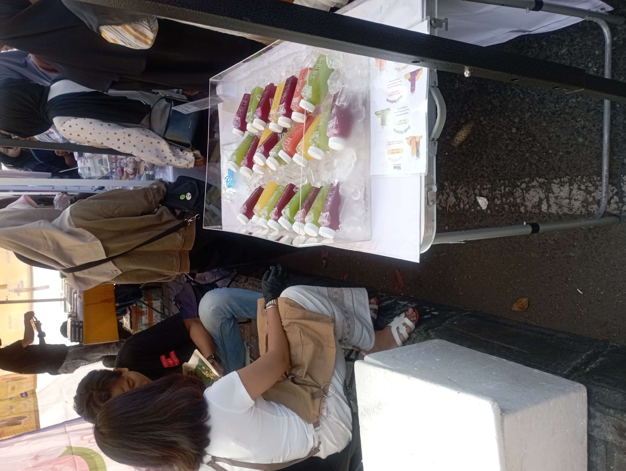 In Depok, Indonesia, a street market scene unfolds. A woman in a white t-shirt and white pants sits on a white styrofoam box, her arm resting on a tan bag. To her left, a man in a black t-shirt and blue jeans leans forward, examining a book. Behind them, various individuals are visible, some wearing headscarves, suggesting a predominantly Muslim population.
The focal point of the image is a clear acrylic container filled with numerous bottles of colorful juices, nestled amongst ice. The juices range in hue from deep purple and vibrant green to sunny yellow and bright orange, indicating a variety of flavors. The container sits on a white table, which also displays printed informational sheets with images of fruits, likely detailing the juice ingredients and benefits.
The ground is paved with asphalt and small stones, showing signs of wear. The overall atmosphere suggests a bustling outdoor market, possibly a food or craft fair, with people interacting and browsing. The presence of a black metal pole on the right side of the frame indicates some form of canopy or display structure, common in street vending setups. The lighting suggests it is daytime, with bright sunlight casting shadows.