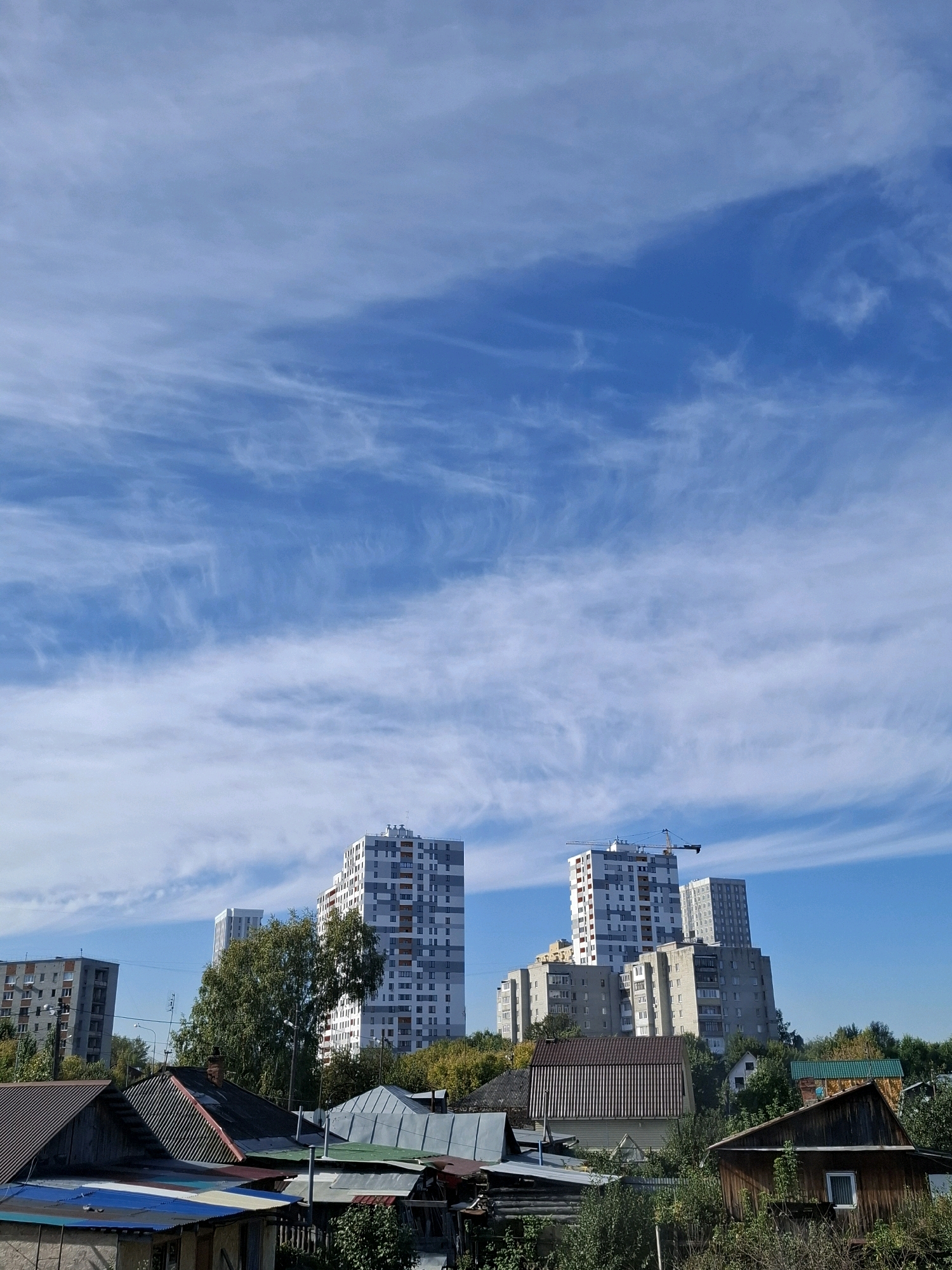 This is a daytime, eye-level, medium shot taken from a residential area looking towards a construction site and older buildings in Tyumen, Russia. The foreground is dominated by the rooftops of low-rise, rustic structures, some made of corrugated metal and wood, indicating a more established or perhaps older part of the city. Behind these, a line of trees separates the older housing from taller, more modern apartment buildings. The tallest of these is a multi-story residential building with a light gray and white facade, featuring numerous windows. A construction crane is visible extending from the top of this building, suggesting ongoing development. To the left of this prominent building, another, slightly shorter apartment building with a gray facade is visible. Further to the left, a lower, concrete apartment block is also present. The sky above is a clear, bright blue, with thin, wispy cirrus clouds streaking across it. The overall impression is one of urban development where newer, taller constructions are being built amidst older residential areas.