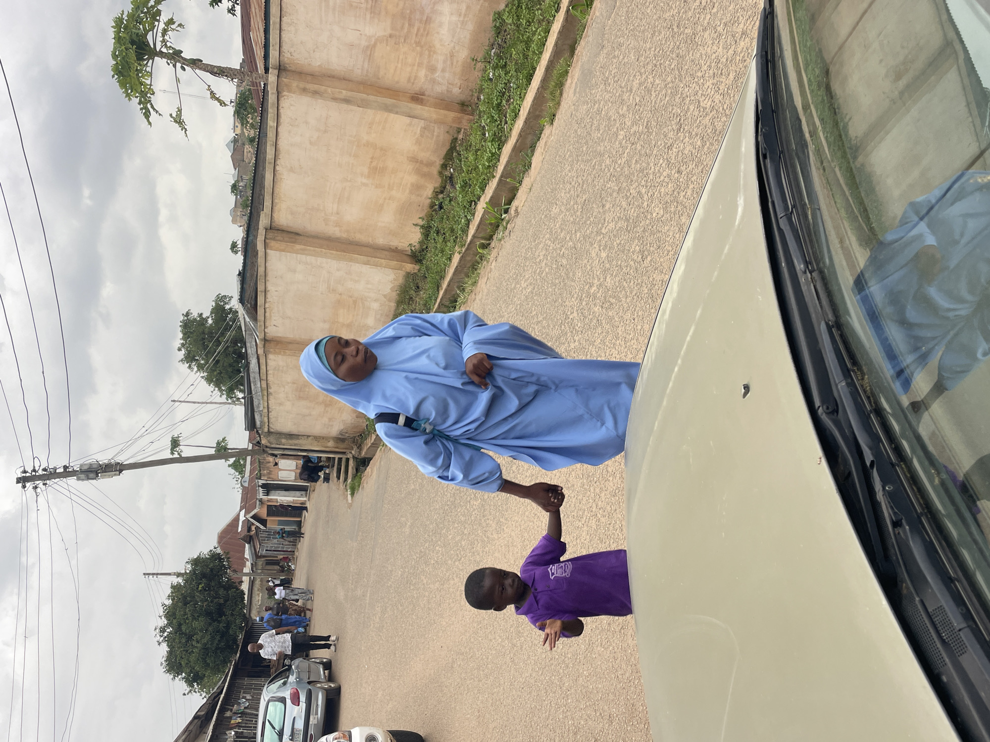 A medium-shot shows a woman in a light blue hijab and a young boy in a purple polo shirt walking hand-in-hand across a paved street in Jos, Nigeria. The woman, who is on the right, appears to be looking down and slightly to her left, while the boy is looking towards the camera with his left arm extended. In the foreground, the hood and windshield wipers of a light-colored car are visible, suggesting the photo was taken from inside another vehicle. The background depicts a street scene with buildings, trees, and utility poles. The sky is overcast. The overall impression is a candid moment captured in an urban environment.